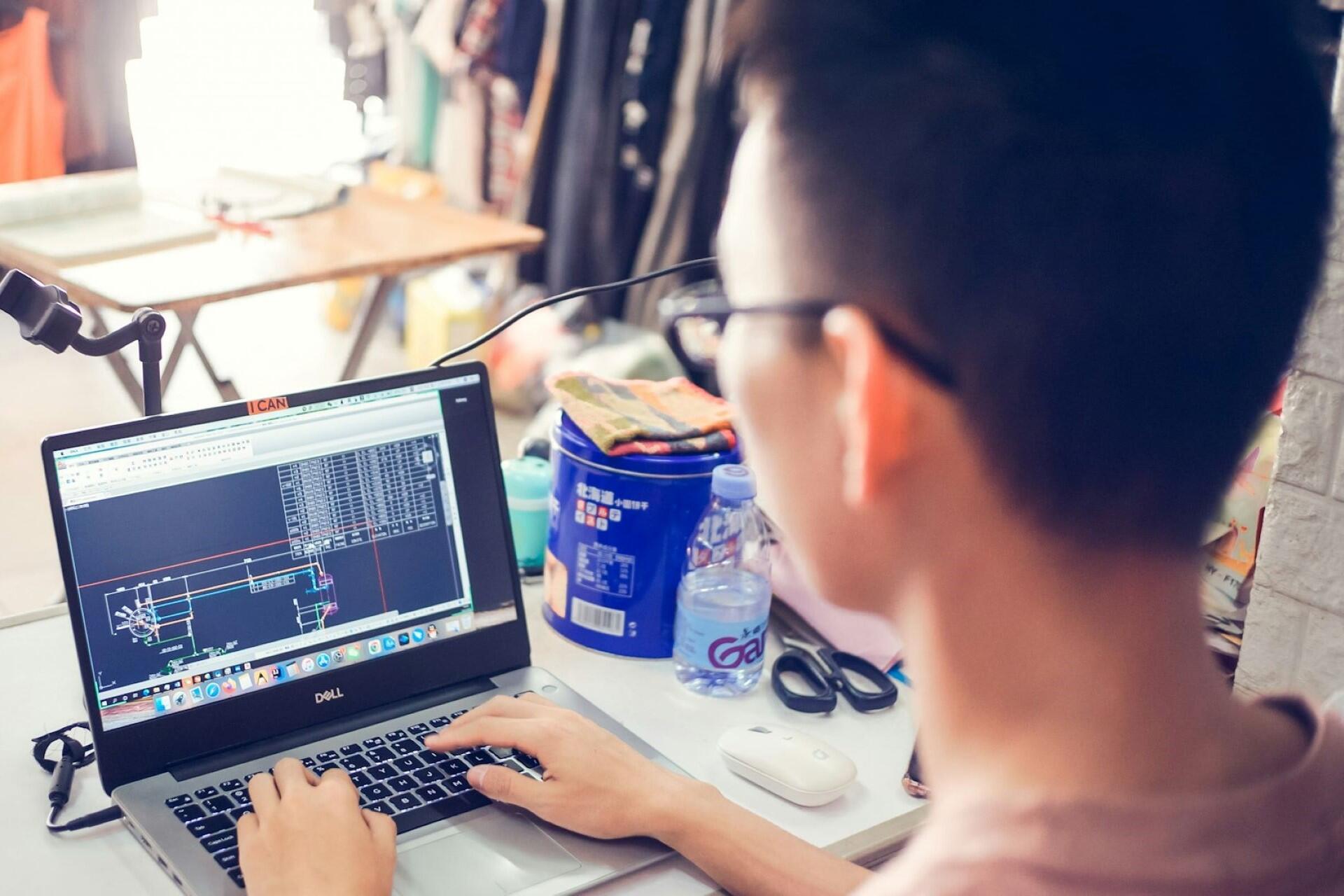 Person wearing glasses using a laptop displaying AutoCAD drafting interface while working at a desk with tools and stationery nearby.