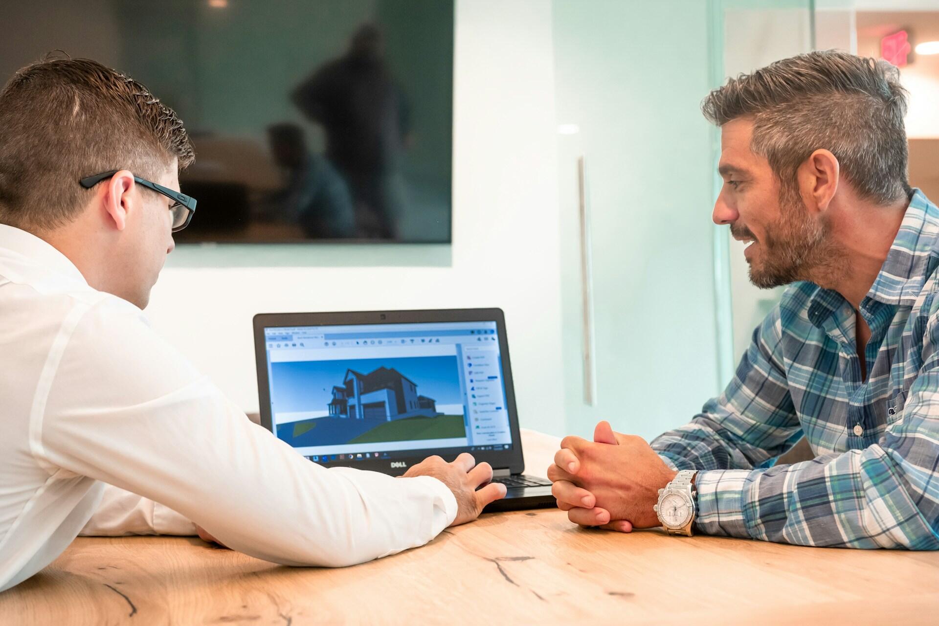 Two men sitting at a wooden table looking at a laptop screen displaying a 3D house model.