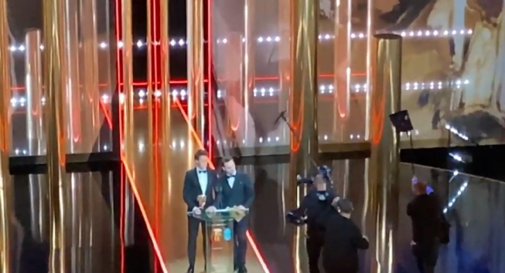 Two men on stage with cameras and boom mikes in front of them. 