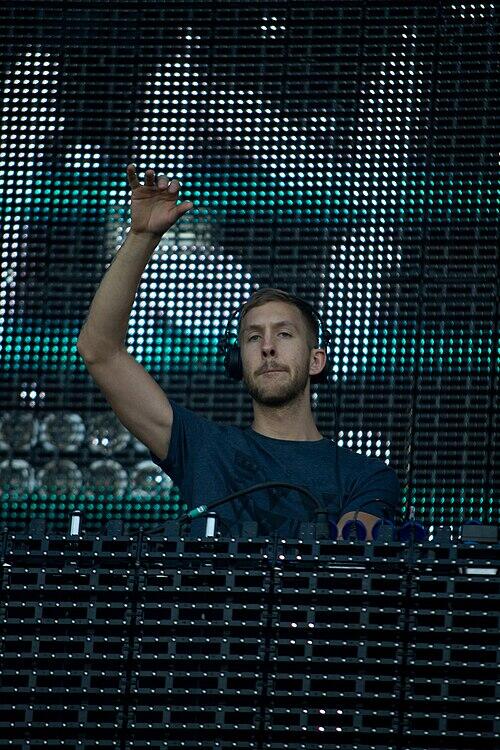 A DJ performs at a live event, gesturing with one hand, behind a mixing console with bright LED screens in the background.