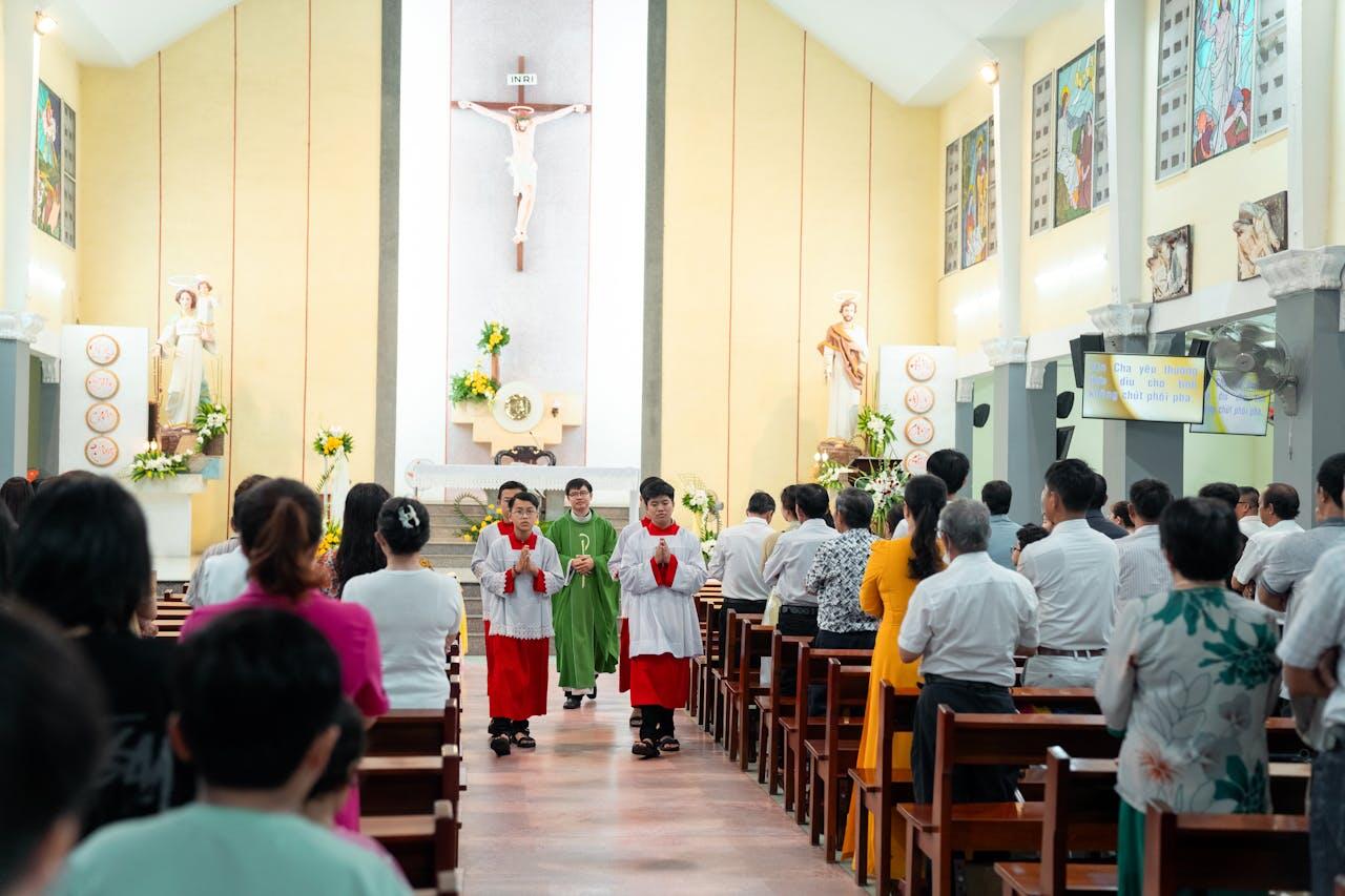 A group of worshipers stand in a church, with altar servers in colorful robes walking towards the altar, surrounded by religious decorations.