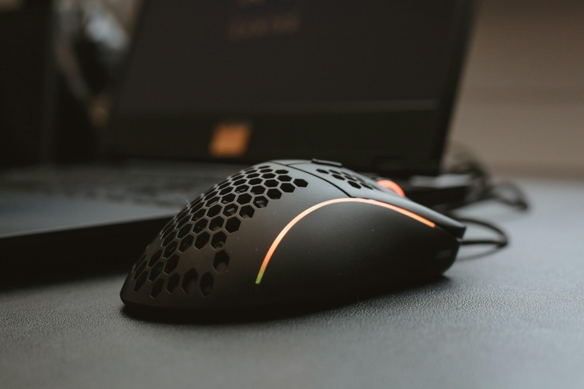 Close-up of a black computer mouse with a honeycomb design and an orange light strip beside a laptop.