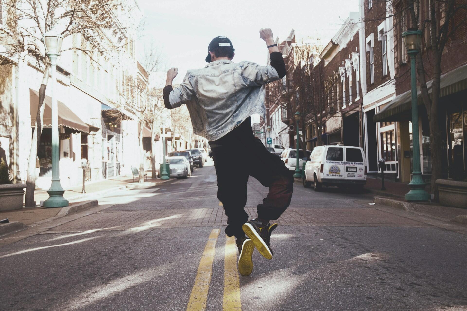 A man wearing casual clothing and a hat dances in the street. 