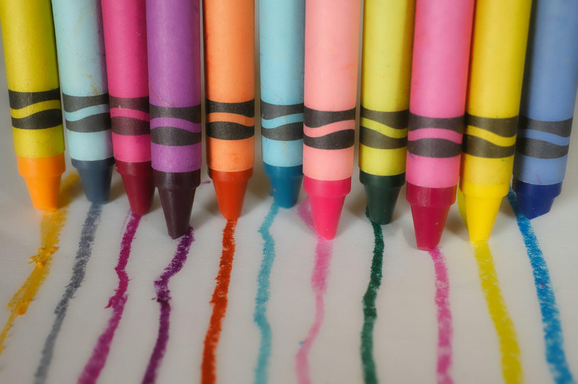 A row of colorful crayons rests on white paper, each leaving vibrant lines of color in various shades.