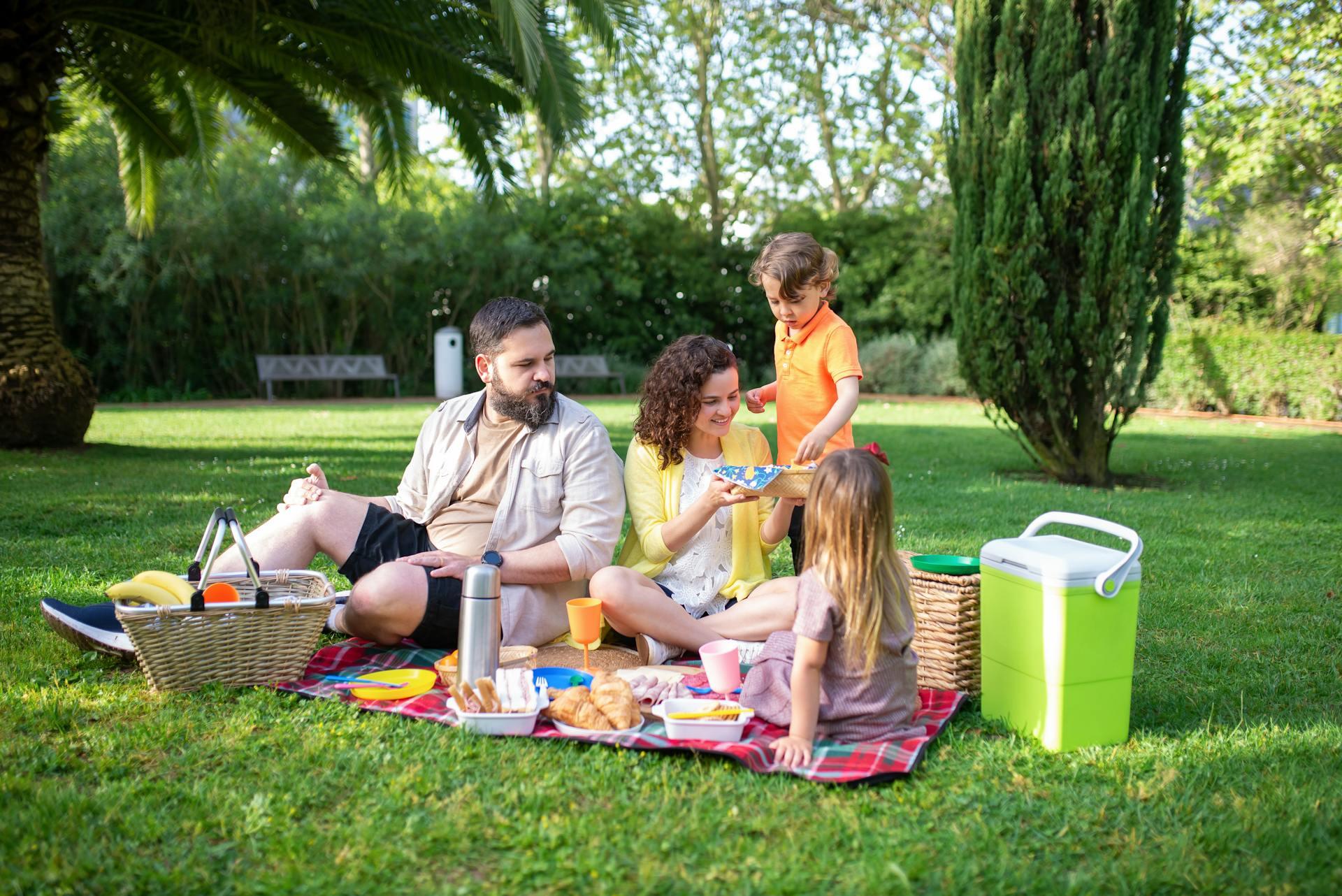 A family enjoys a picnic on a grassy lawn, surrounded by trees, with a spread of food and drinks laid out on a blanket.