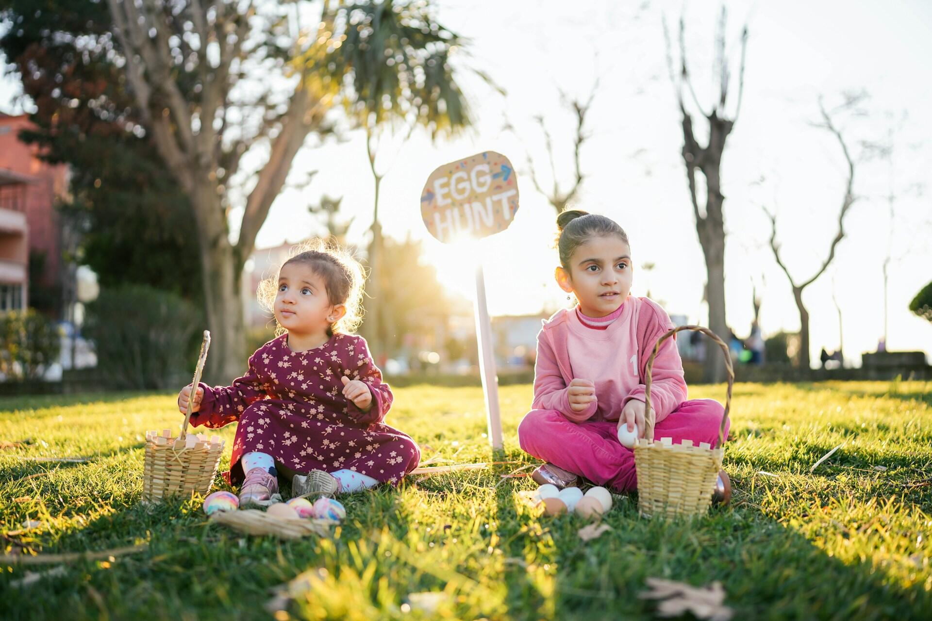 Two children seated on grass, enjoying an egg hunt, with baskets and colorful eggs around them, under the sunlight.