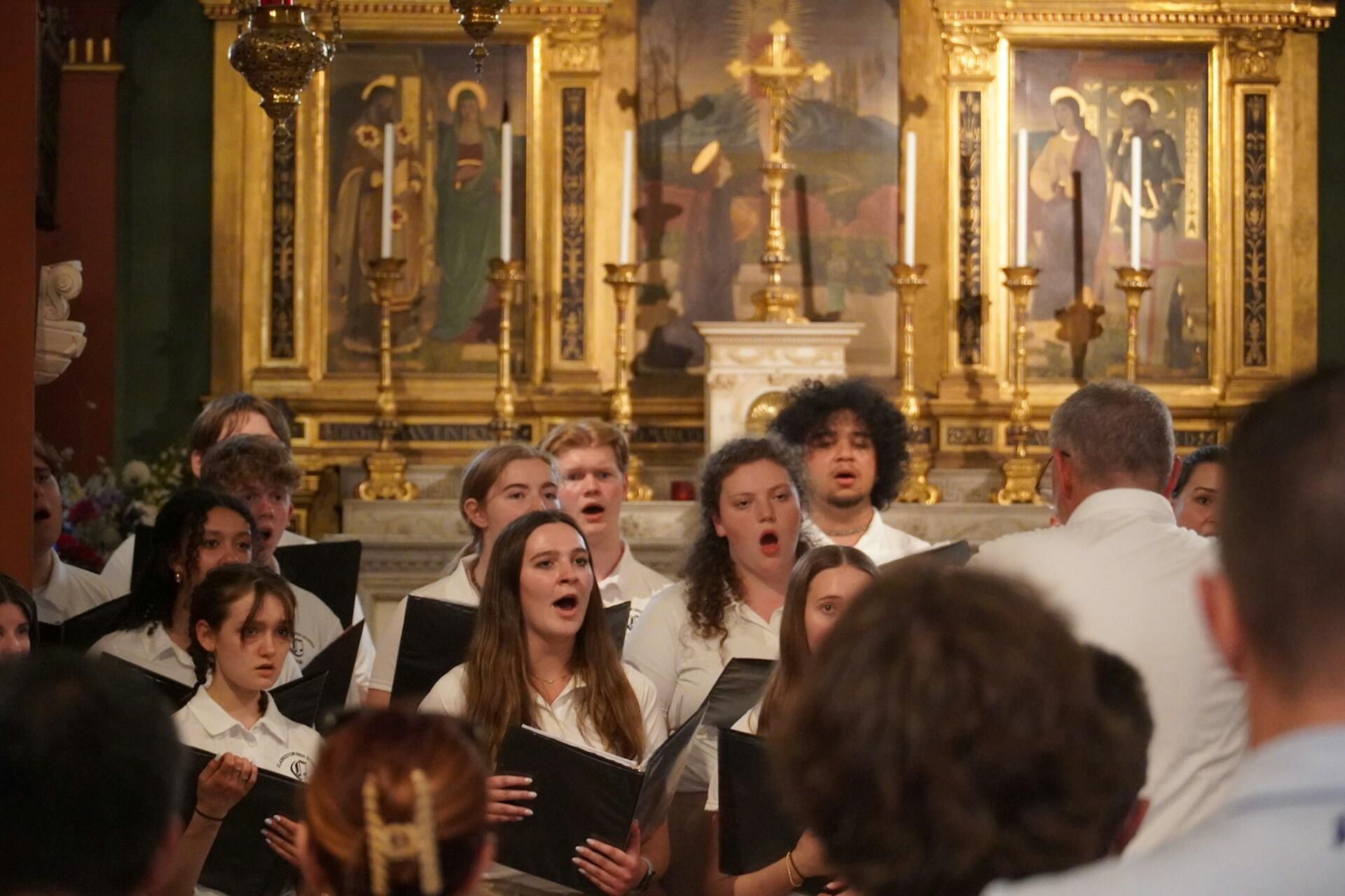 A choir of individuals in white shirts performs in front of an ornate gold altar with lit candles and religious artwork.