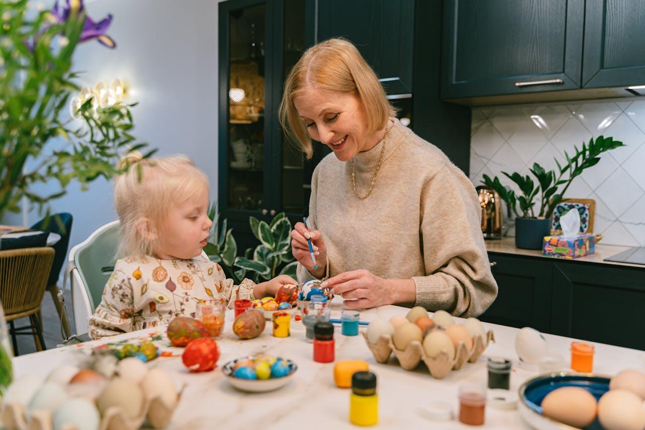 A person and a child are painting colorful Easter eggs at a table filled with paint and decorated eggs in a cozy kitchen.