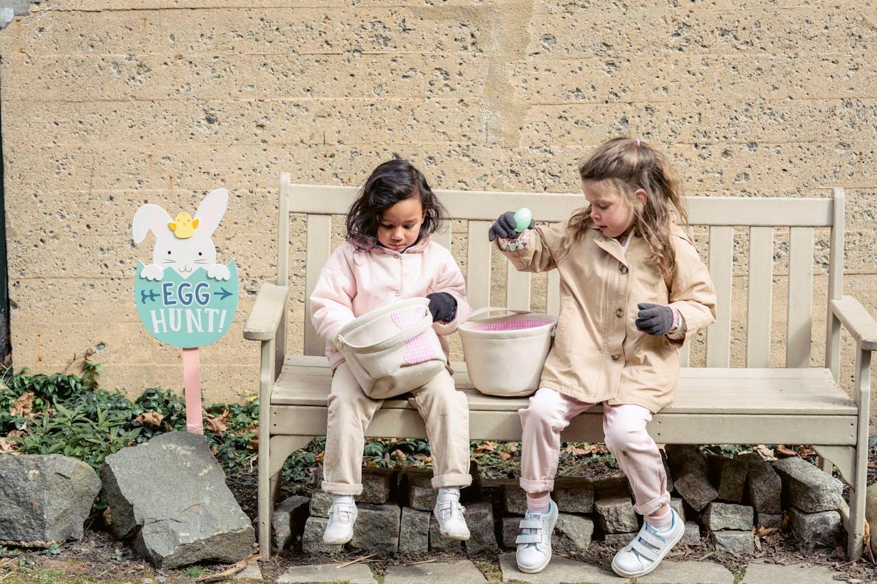 Two children sit on a bench, holding baskets filled with colorful eggs, beside an "Egg Hunt!" sign in a garden setting.