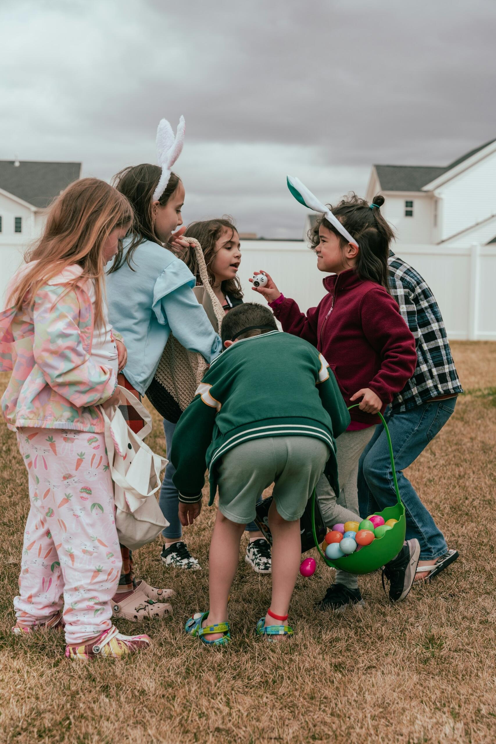 A group of children gather on grass, eagerly searching for colorful Easter eggs, some wearing bunny ears and casual clothing.