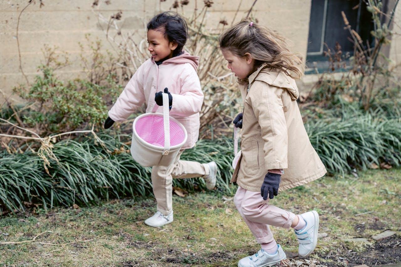 Two children in light jackets and gloves run excitedly through a garden, one carrying a small pink basket.