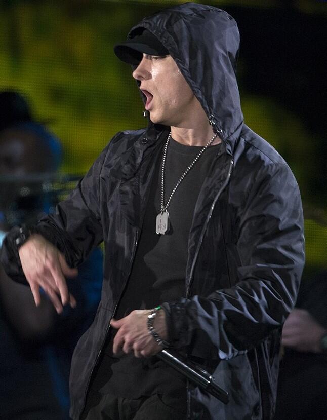 A performer in a black hooded jacket and dog tags gestures while holding a microphone on stage, illuminated by bright lights.