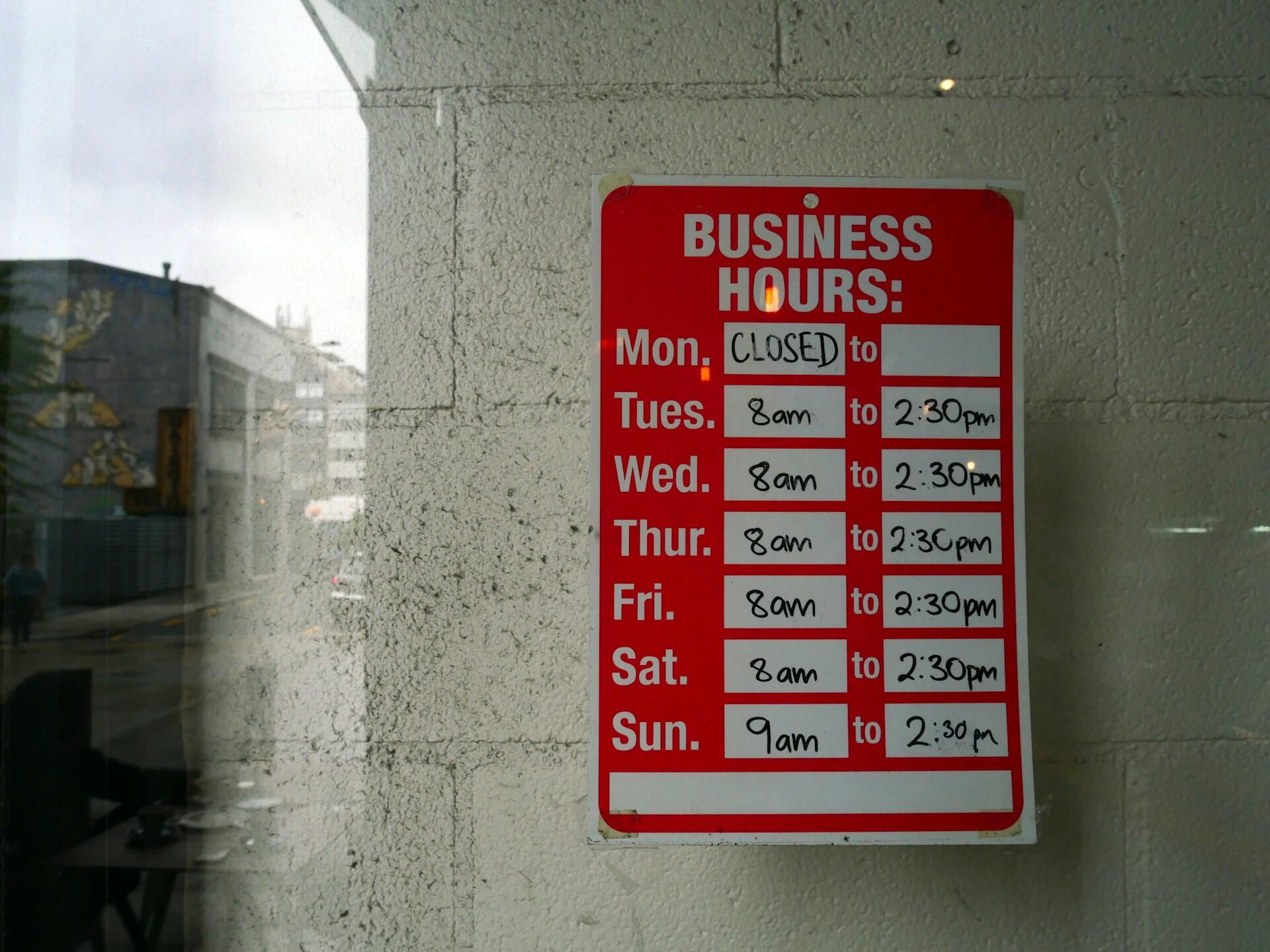 A red sign on a window listing daily operating hours. 