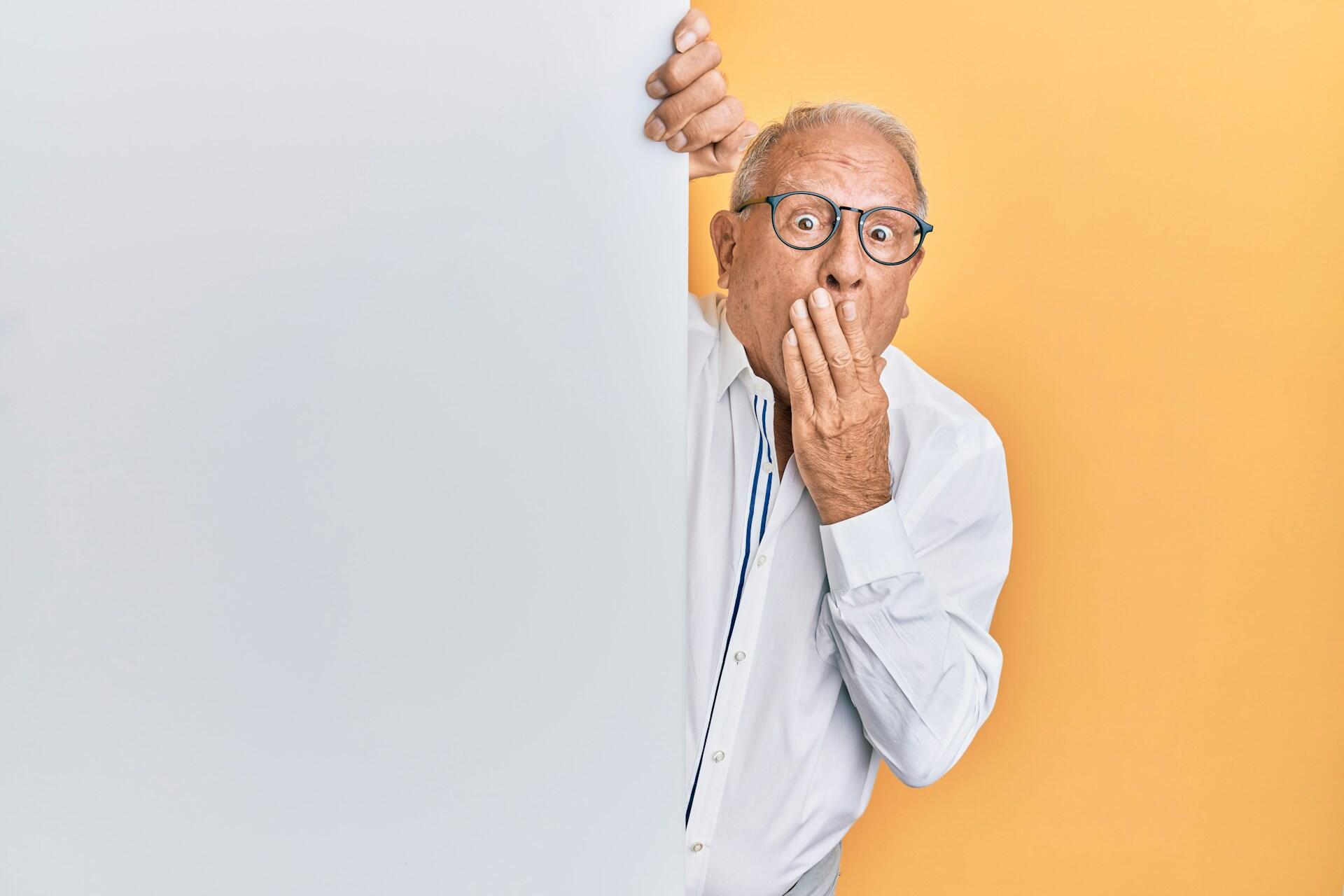 A bald man wearing glasses partly hidden behind a wall holds his hand over his mouth.