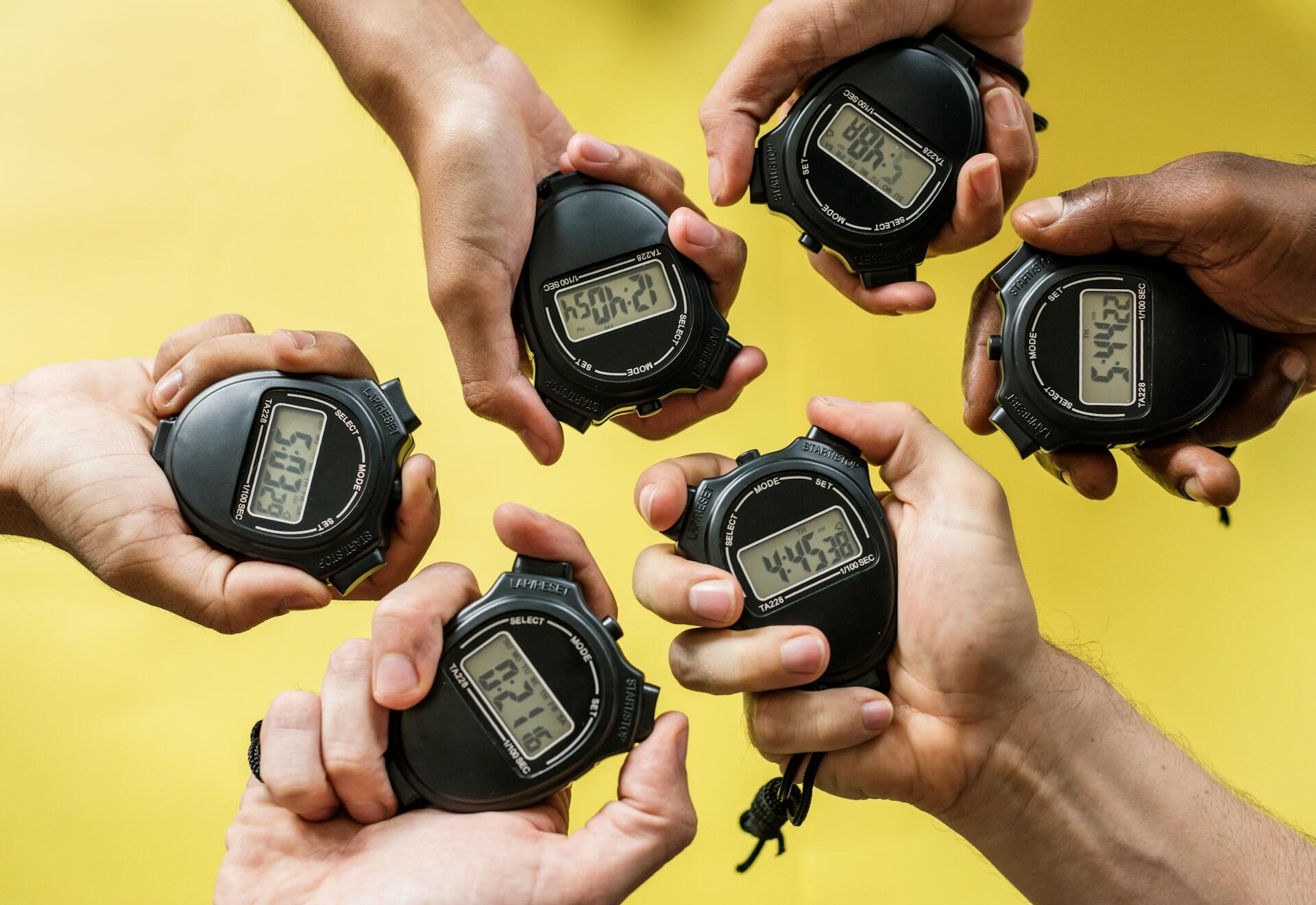 Six hands, each holding a stopwatch showing different times. 