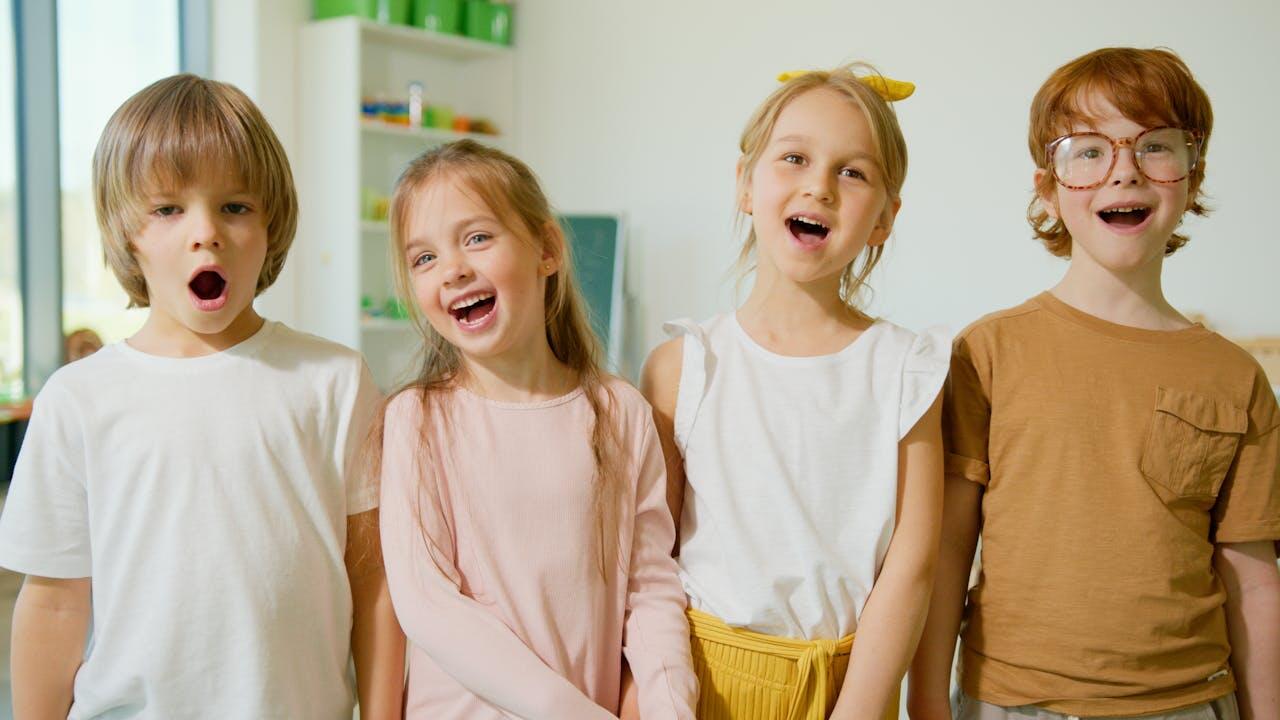 Four children stand side by side in a bright room, each wearing casual attire, in front of shelves filled with colorful items.