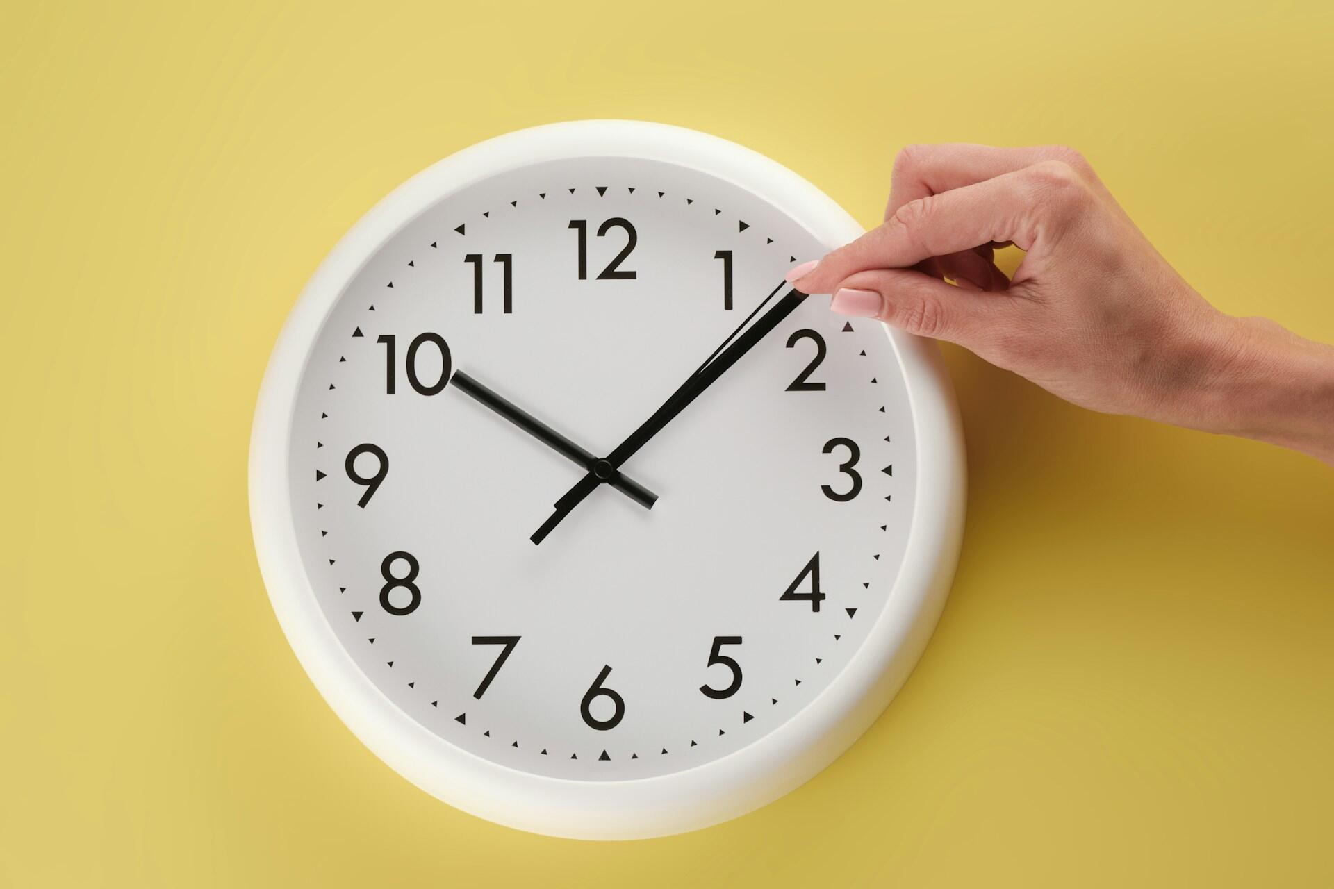 A person's hand adjusting a white clock with black hands on a yellow wall.