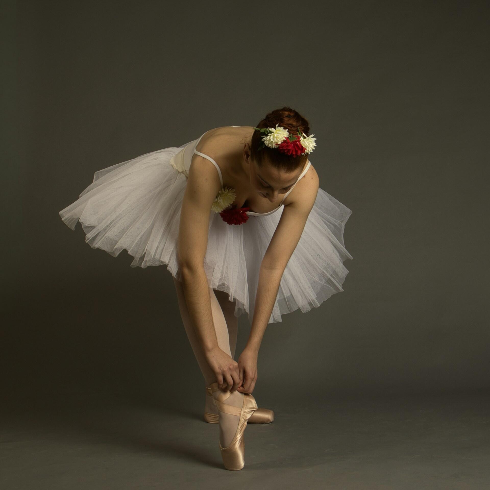 A ballerina in a white tutu and flowered headband adjusts her ballet shoes. 