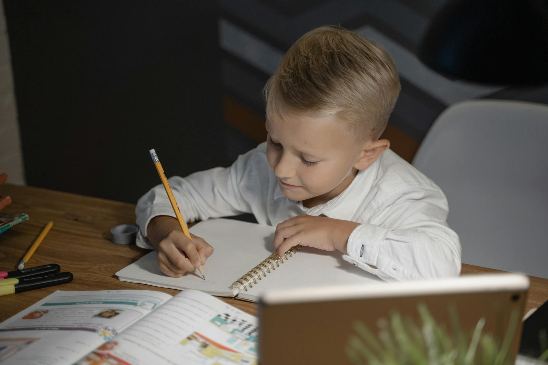 A child in a white shirt writes in a notebook at a wooden table surrounded by colorful pens and books.
