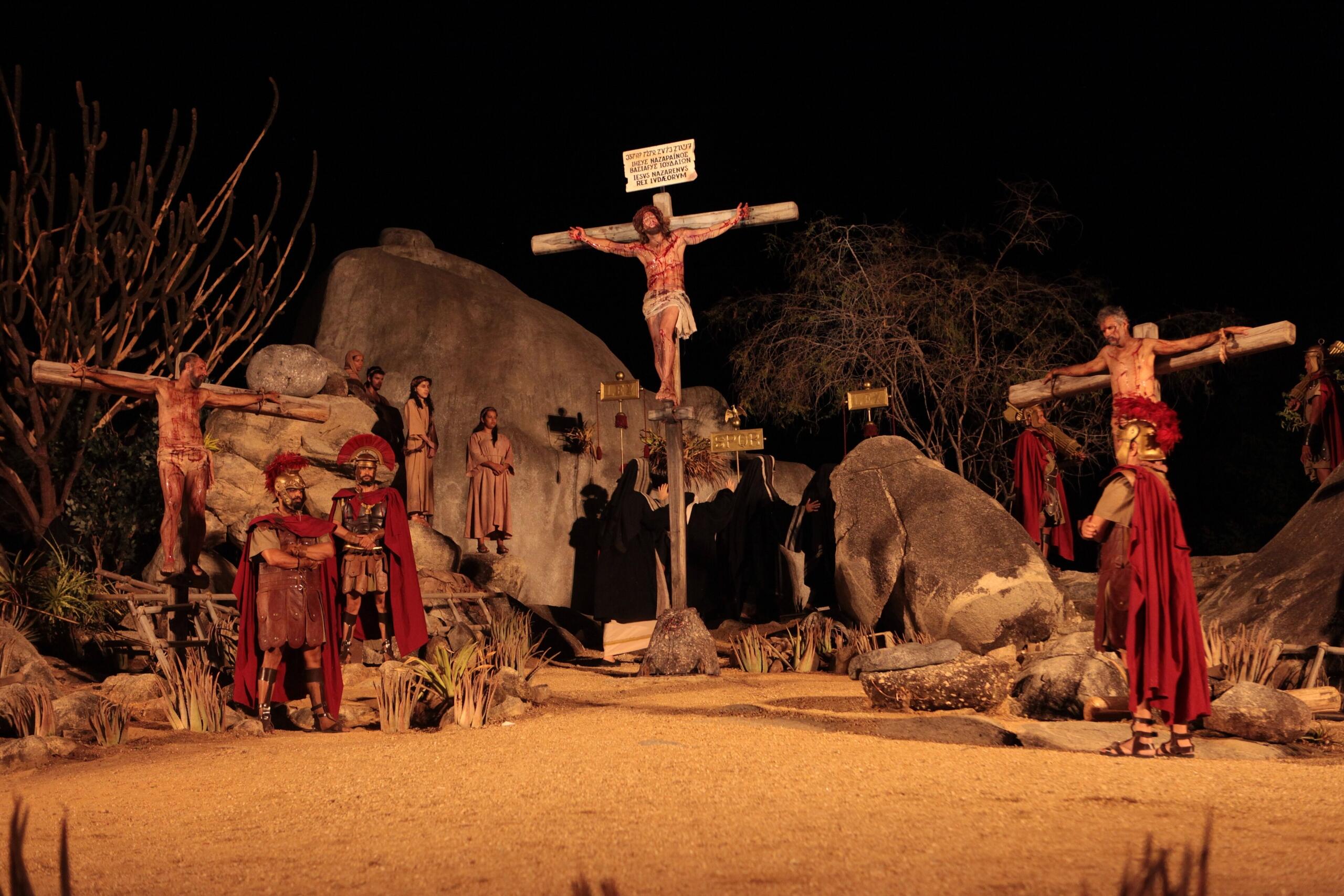 Dramatic night scene depicting a crucifixion reenactment, with figures in historical costumes surrounded by rocks and sparse vegetation.