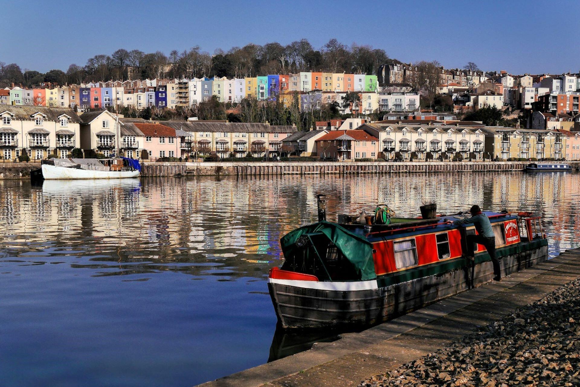 Boats moored on the water in Bristol Harbour with colourful houses lining the hillside in the background.
