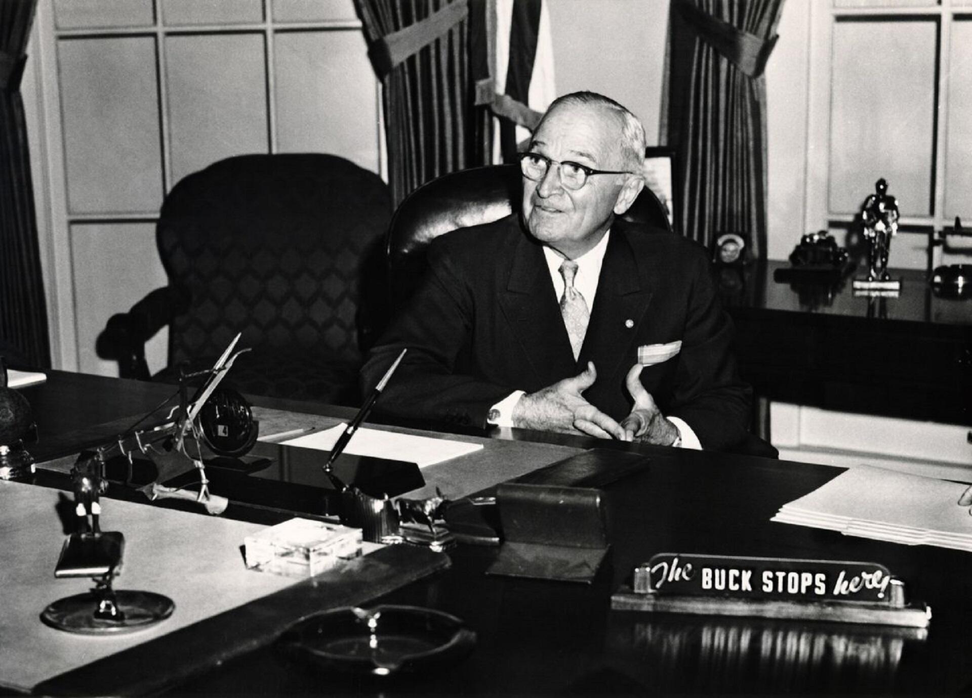 A man sitting behind a desk which features a sign saying 'the buck stops here'. 