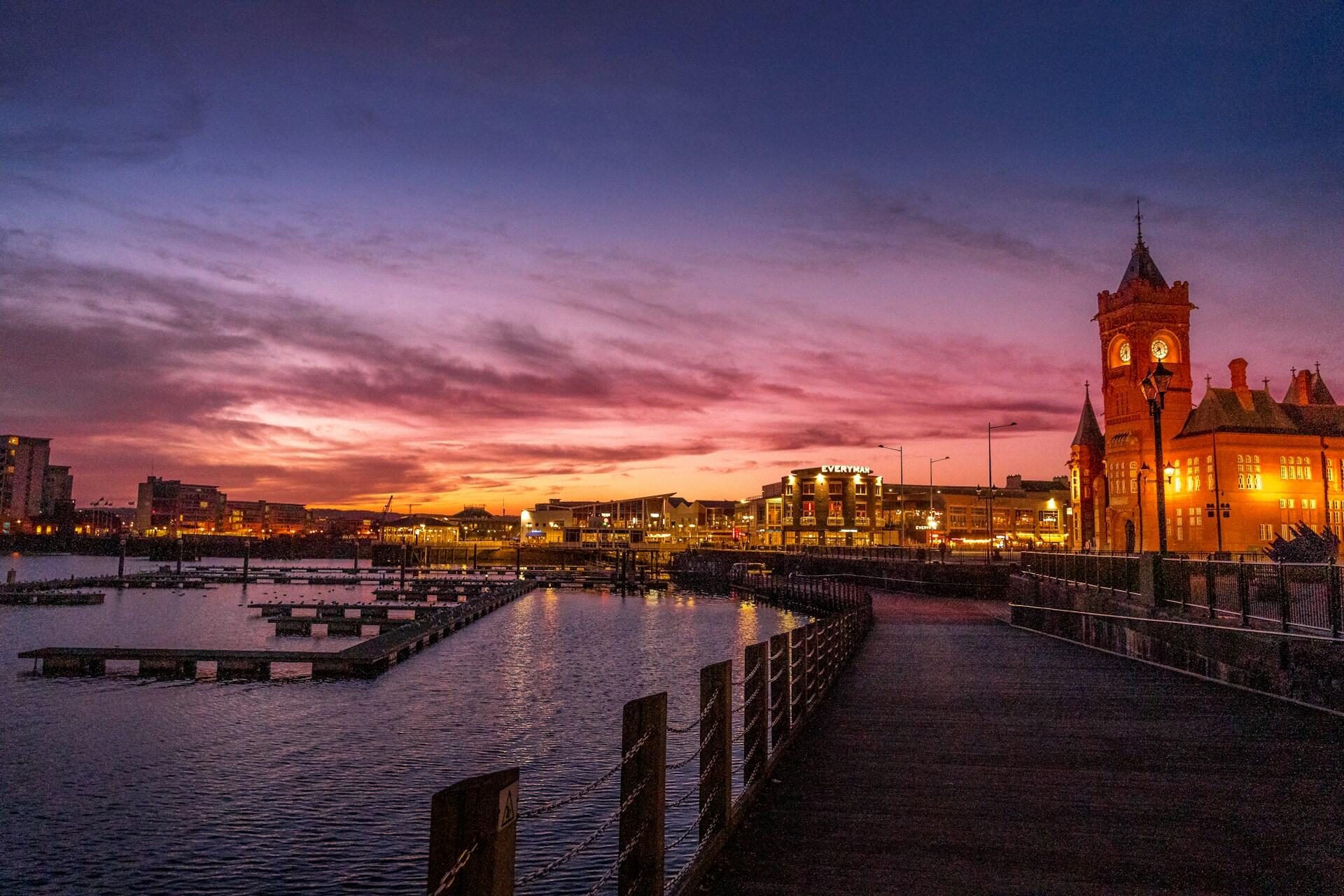 Cardiff Bay waterfront at sunset with buildings, boats, and a lit clock tower
