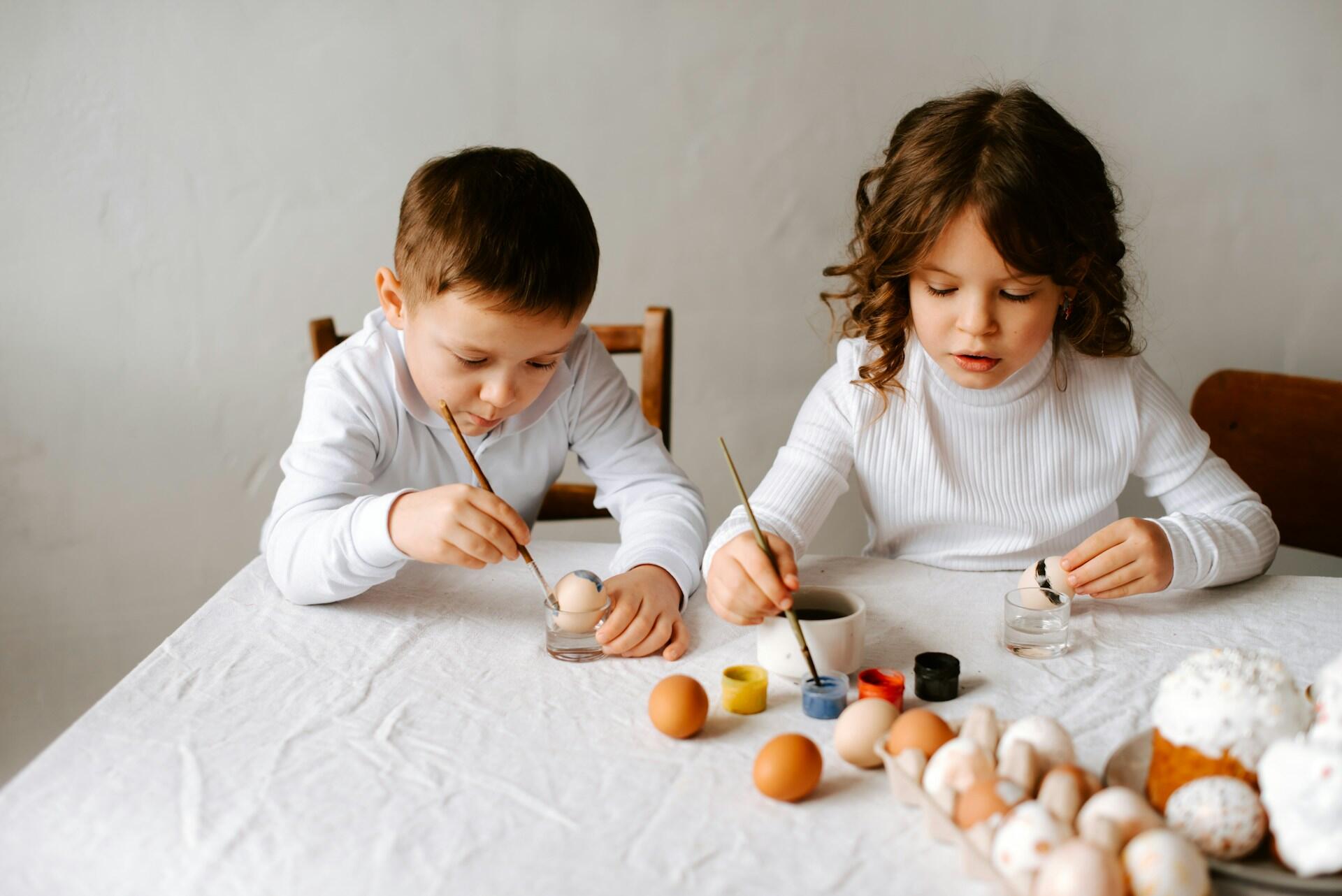 Two children in white sweaters paint eggs at a table covered with a cloth, surrounded by brushes and paint containers.