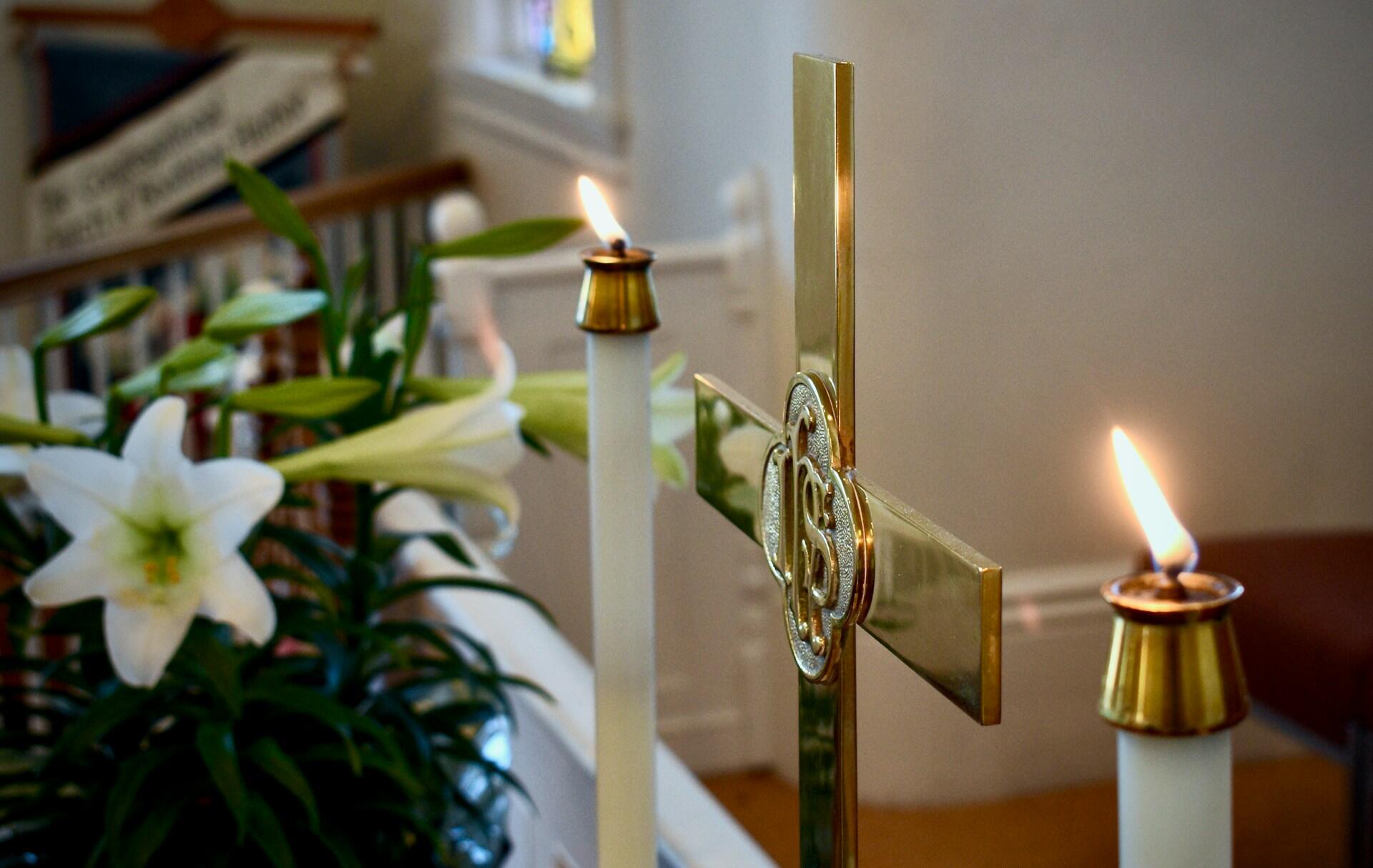 Golden cross with large candles burning on either side, on a table in a church. Easter lilies bloom in the background.