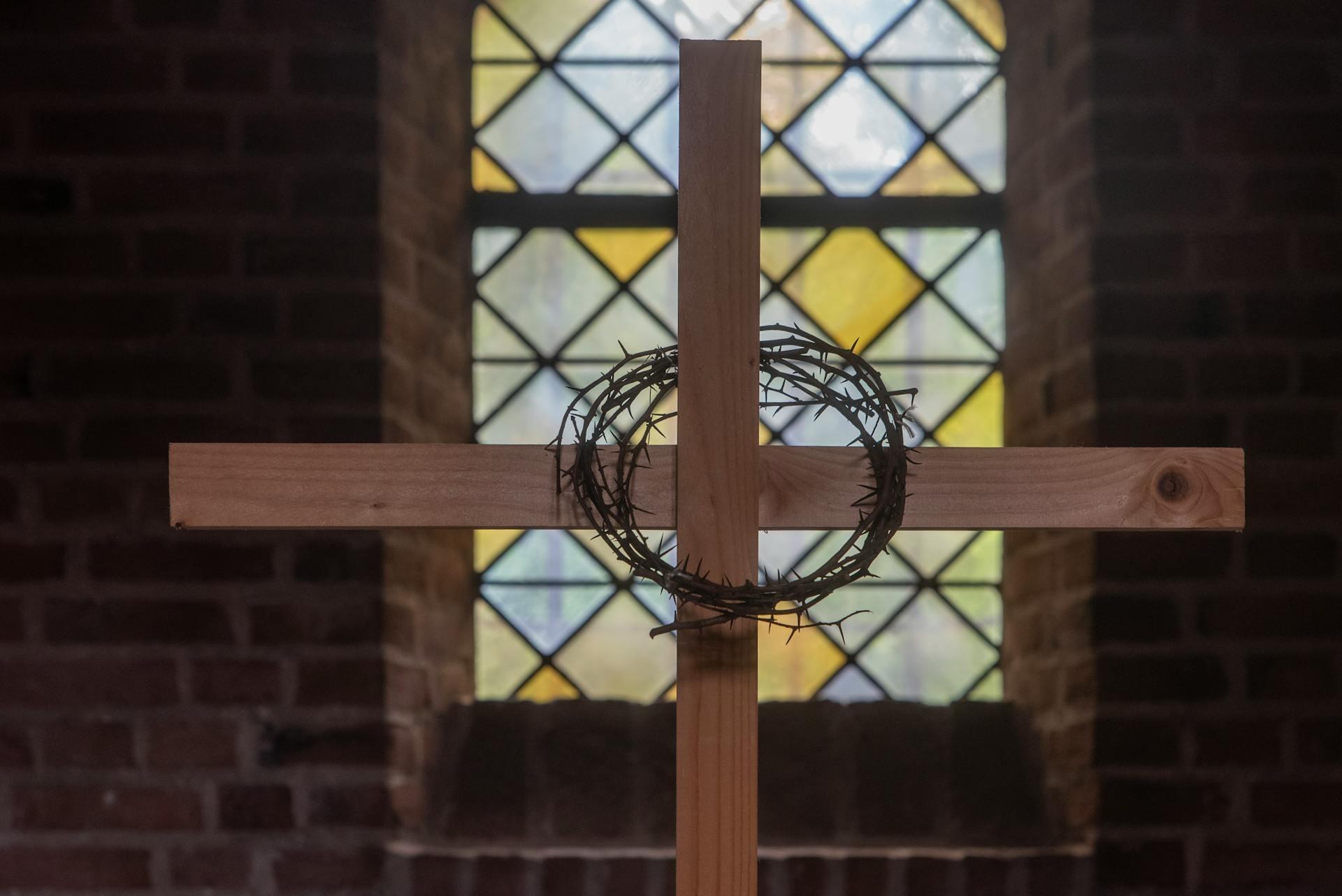 Wooden cross with a crown of thorns hanging on it, set against a stained glass window background in a dimly lit brick church.