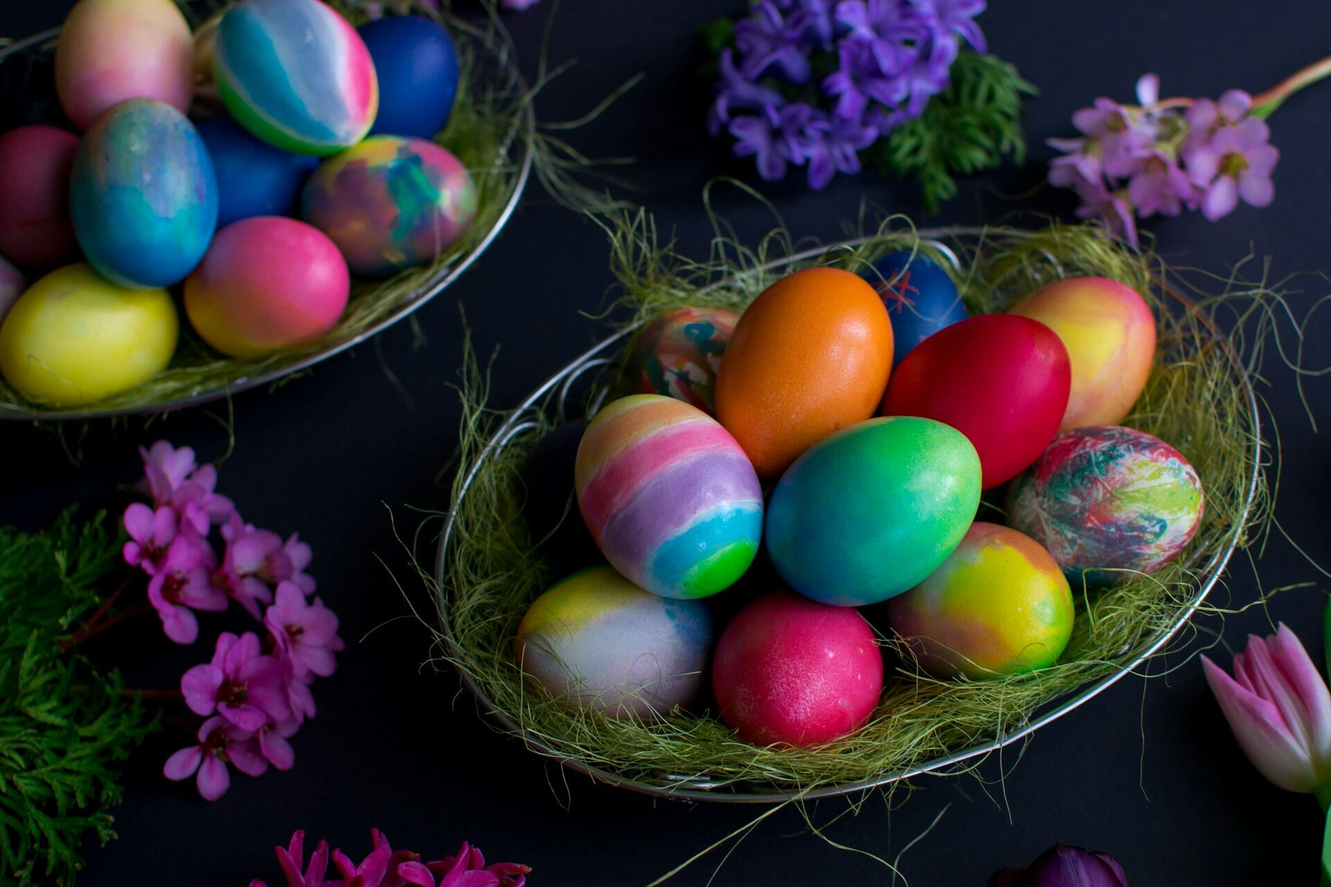 A collection of vibrant, decorated Easter eggs in a nest with colorful flowers and greenery on a dark background.