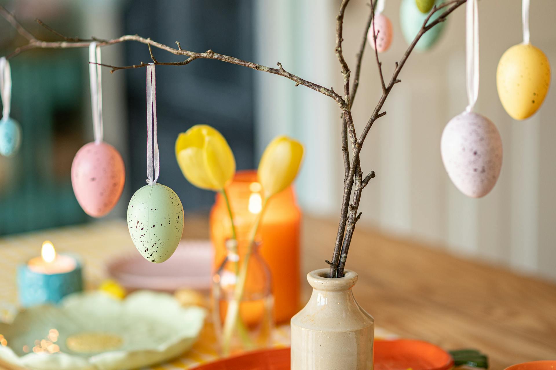 A decorative branch with pastel-colored hanging eggs, surrounded by tulips, candles, and colorful tableware, set for a festive occasion.
