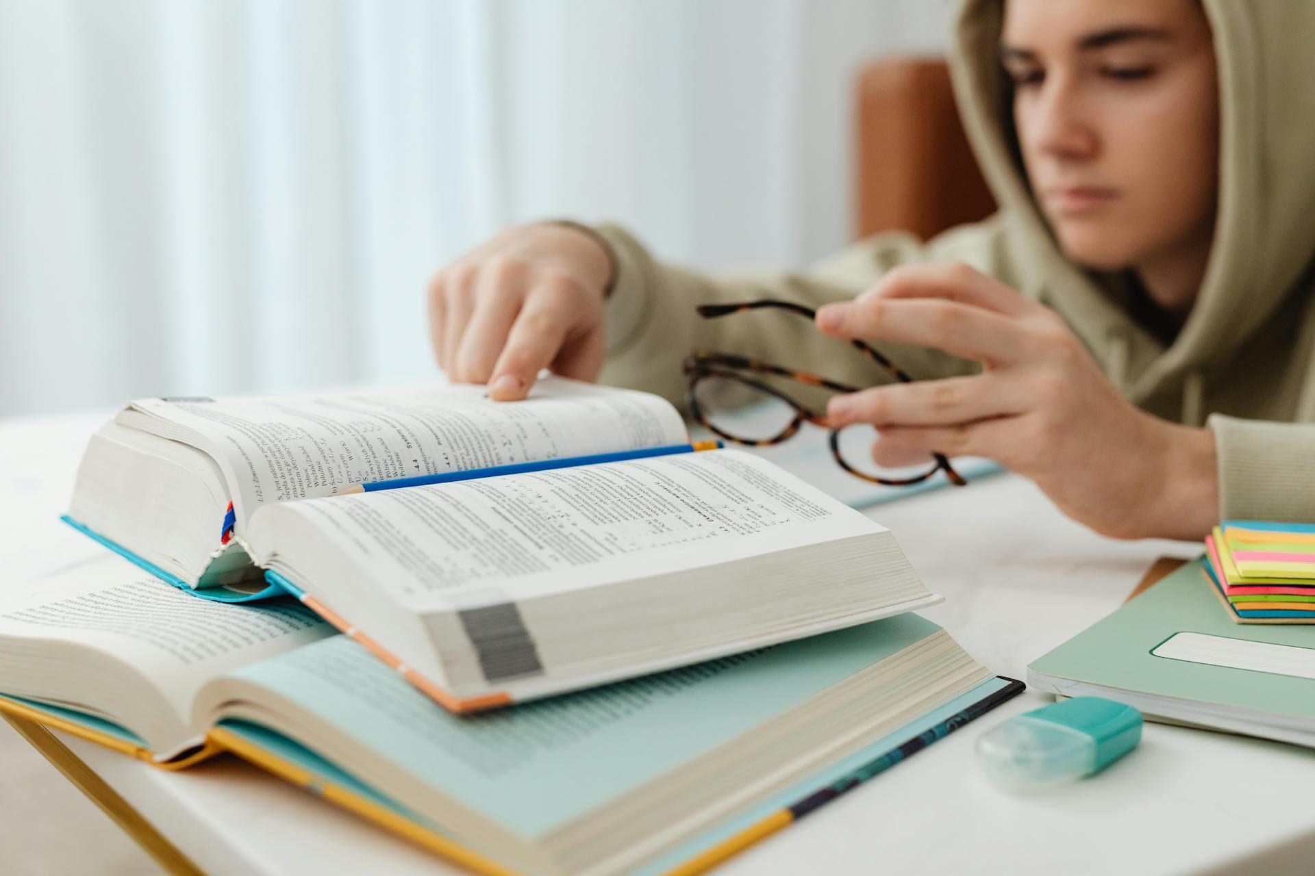 Young person with glasses in hands leaning over and reading from school book