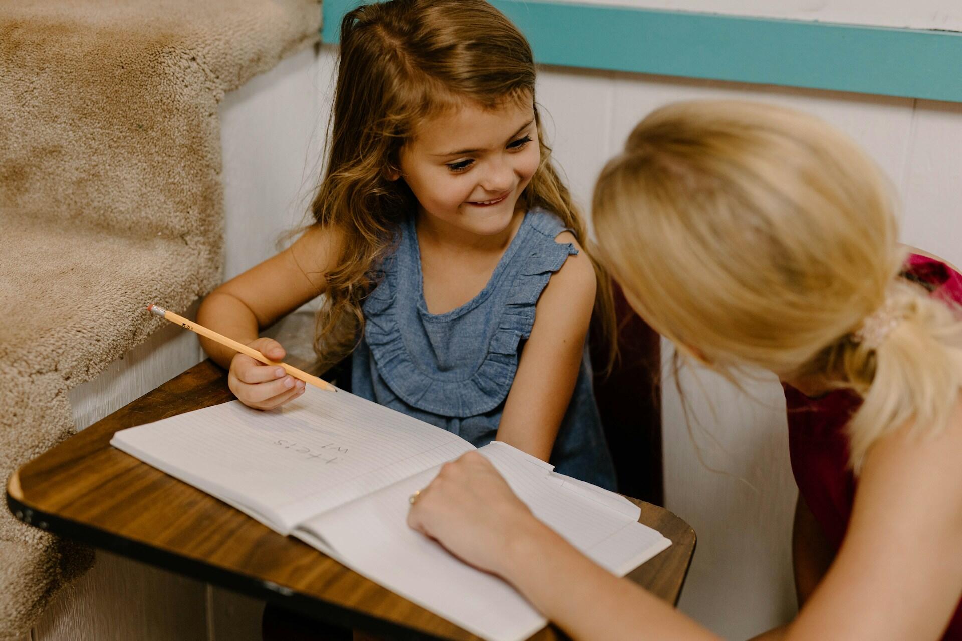 An adult kneels by a seated student who's writing in a notebook.