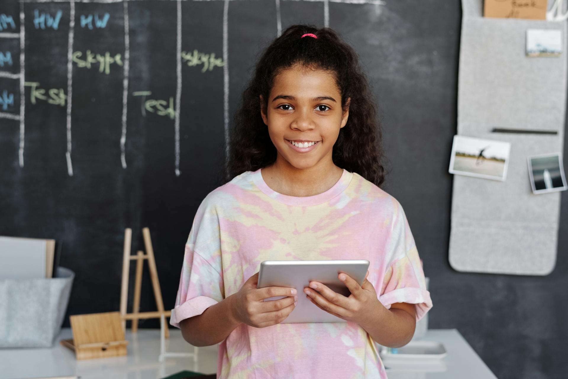 Image of a girl standing in front of a blackboard holding a tablet while smiling into the camera.