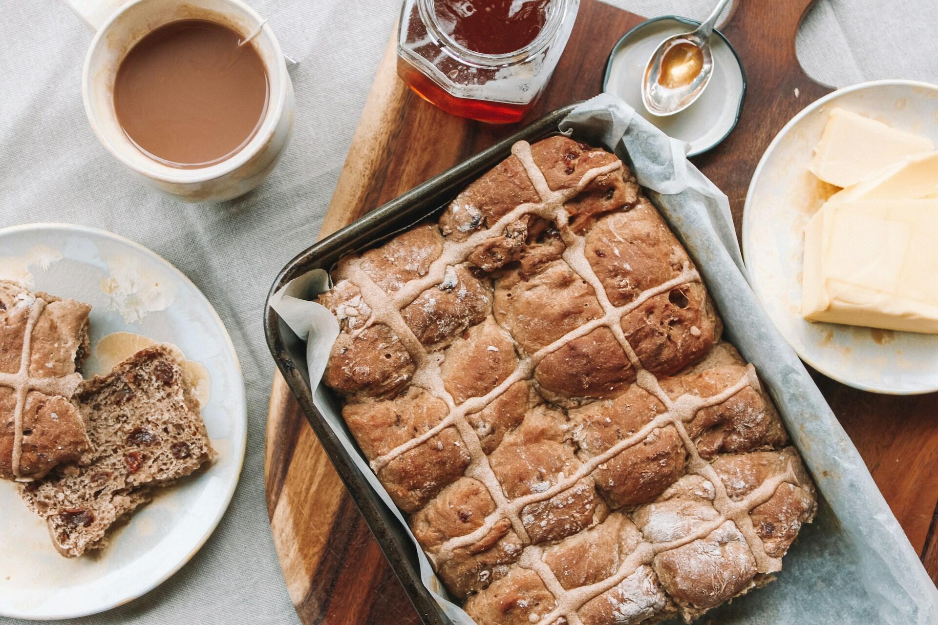 A tray of freshly baked hot cross buns, sliced and served with butter, jam, and a cup of tea on a wooden table.