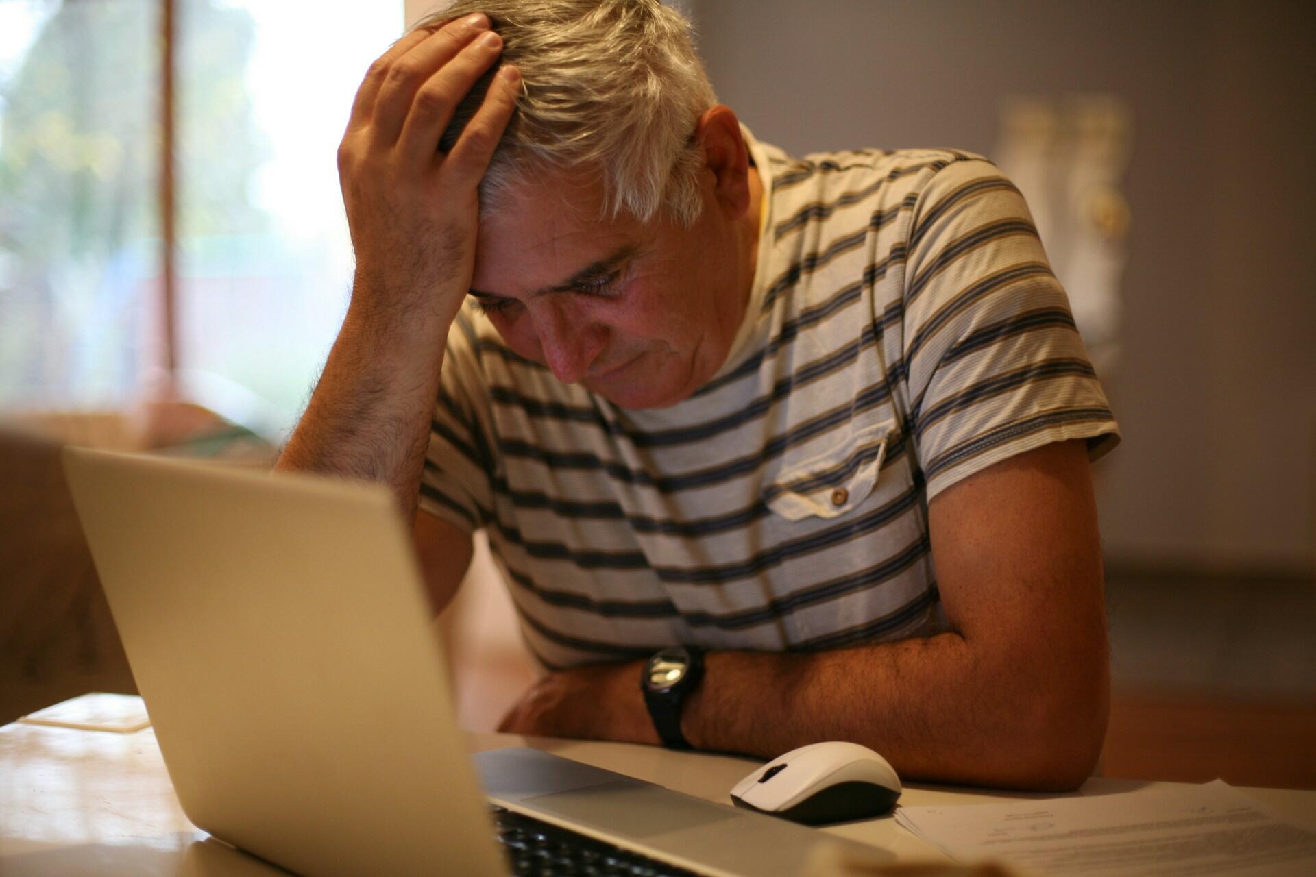 A man wearing a striped shirt holds his head in his hand in front of a computer. 