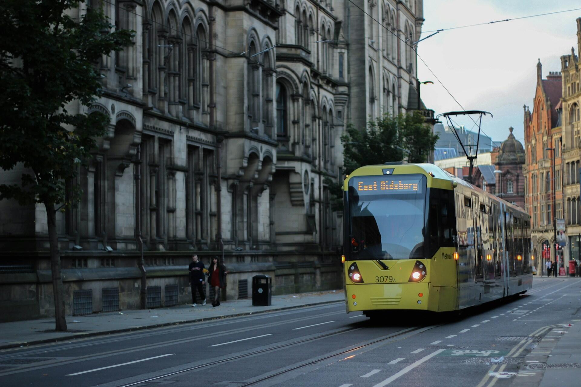 Yellow Metrolink tram travelling through a street in Manchester city centre beside historic buildings