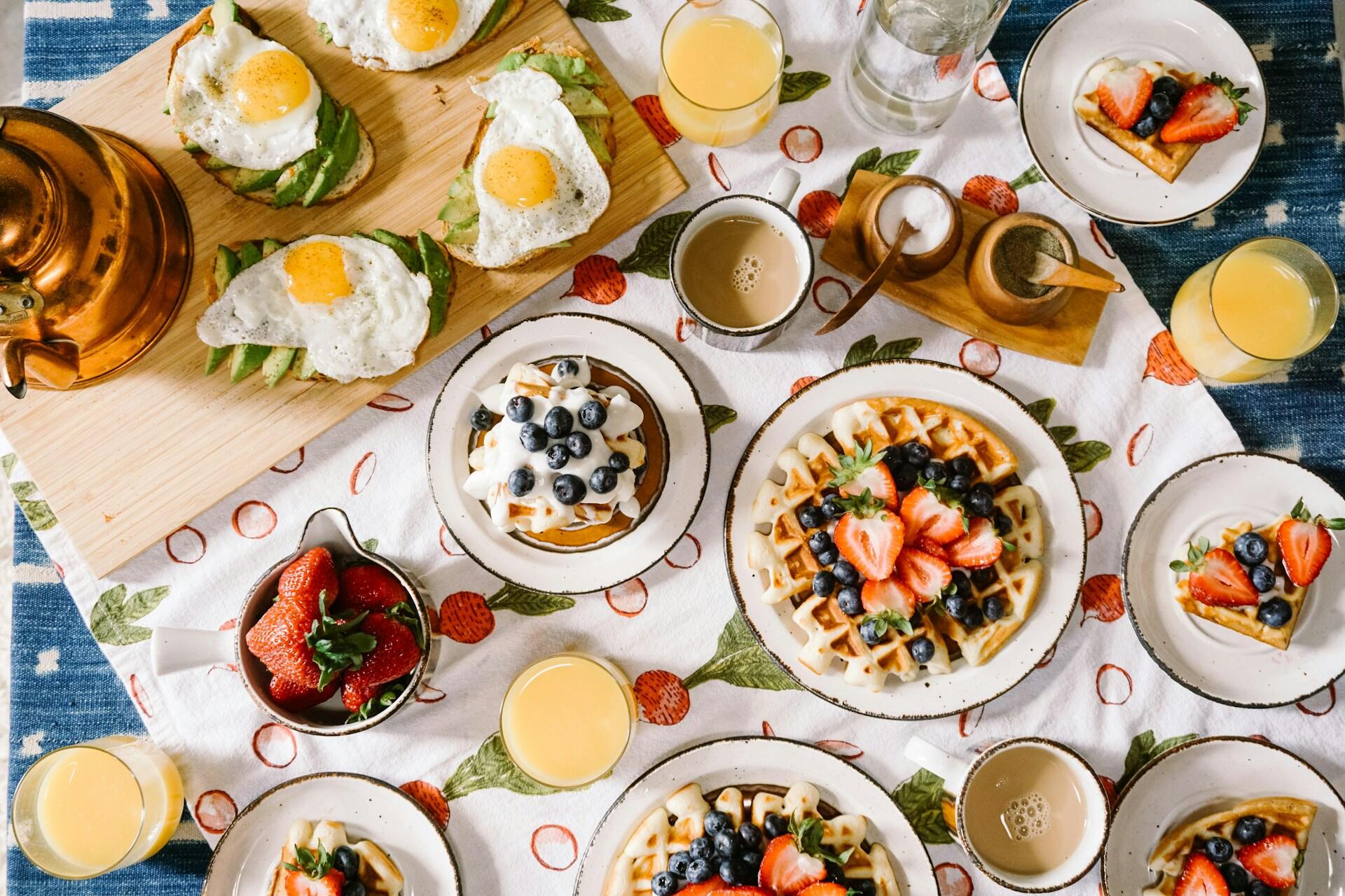 A table loaded with various dishes, including waffles, fruit, and egg toast.