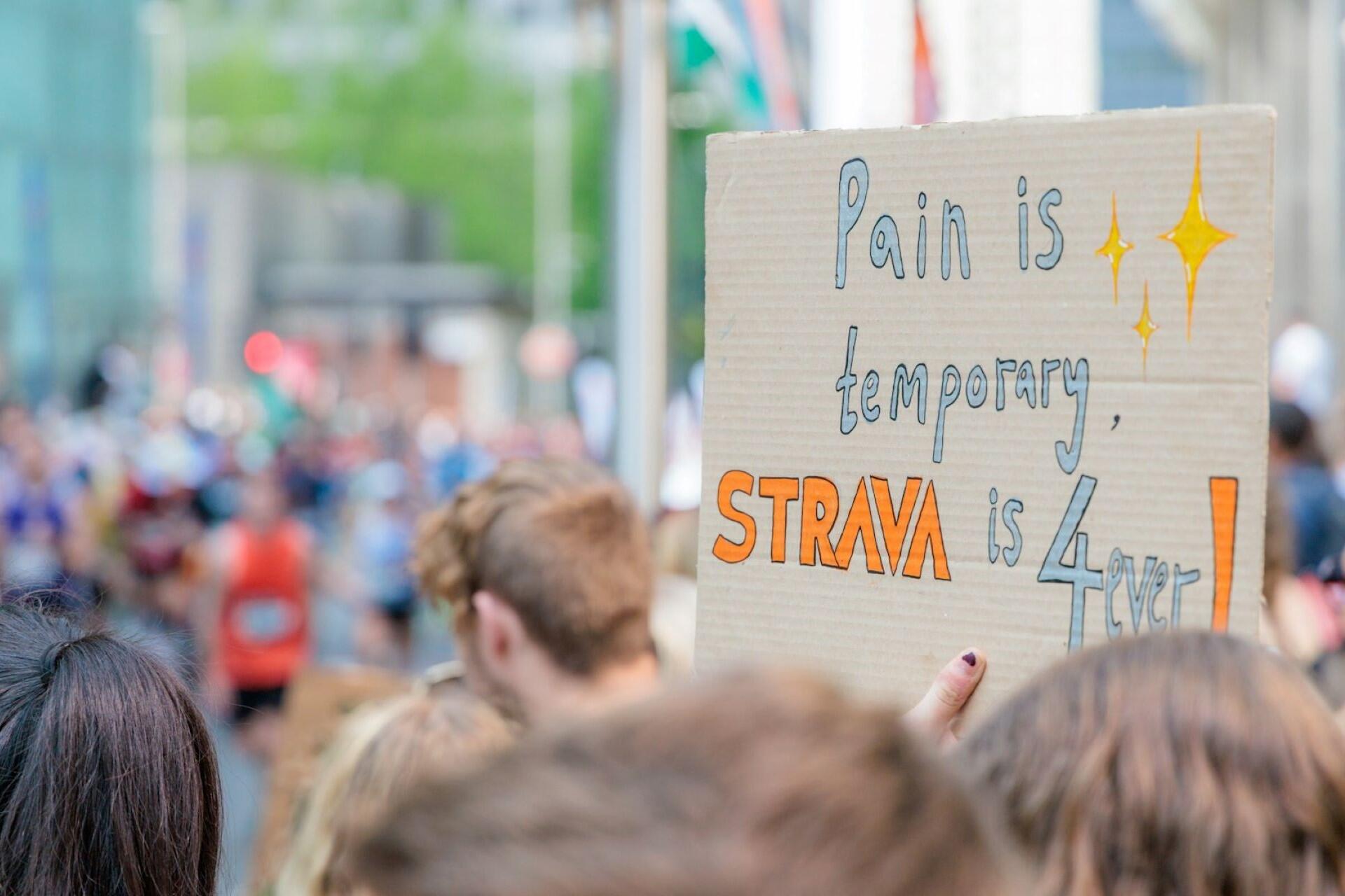 Person in a crowd holding a cardboard sign that reads “Pain is temporary, Strava is forever” during a marathon race.