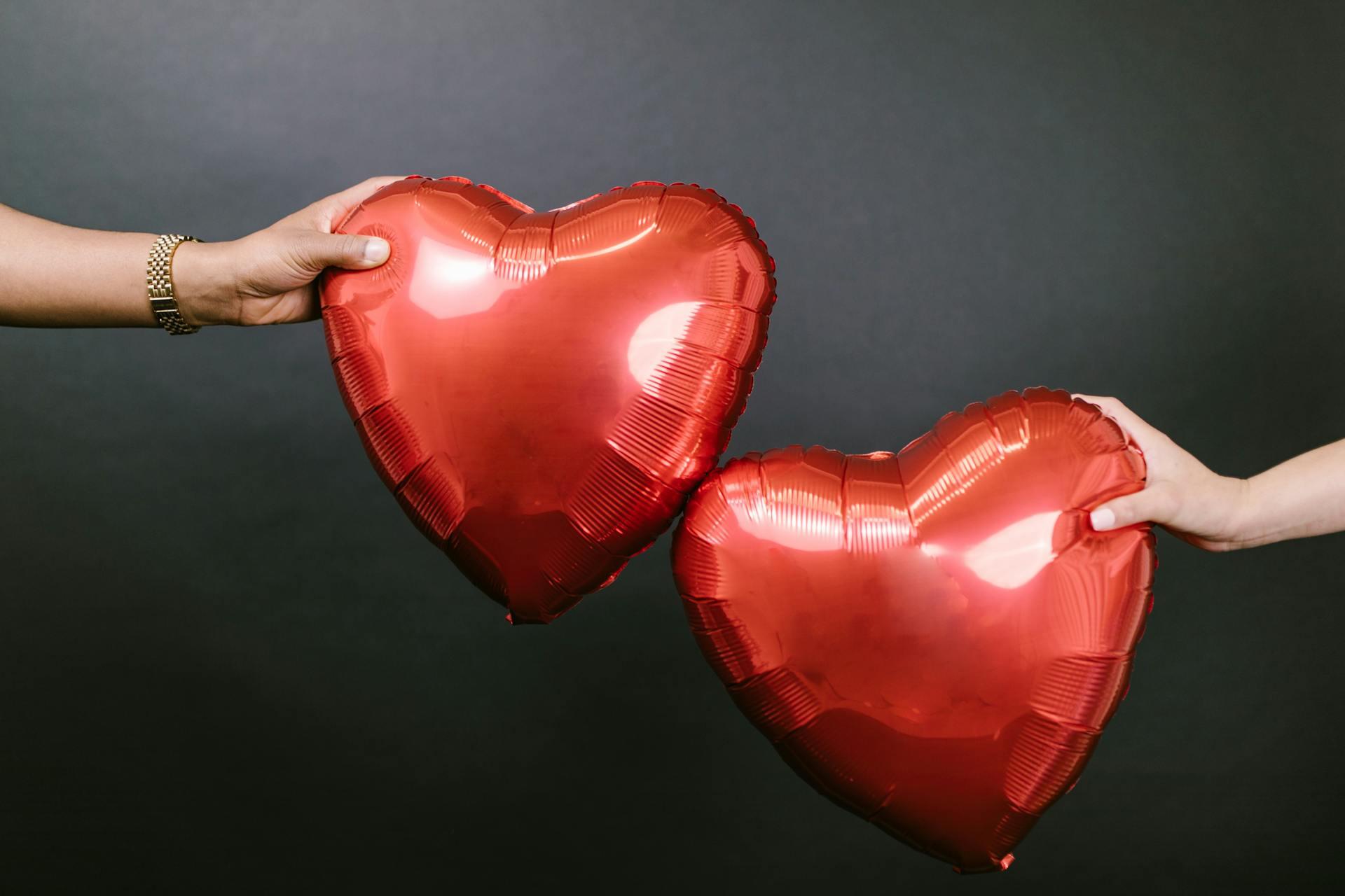 Two hands hold shiny red heart-shaped balloons against a dark background, symbolizing love and connection.