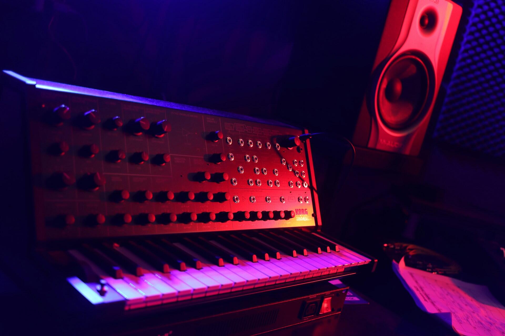 Electronic synthesizer and studio monitor speaker in a dimly lit music production studio.