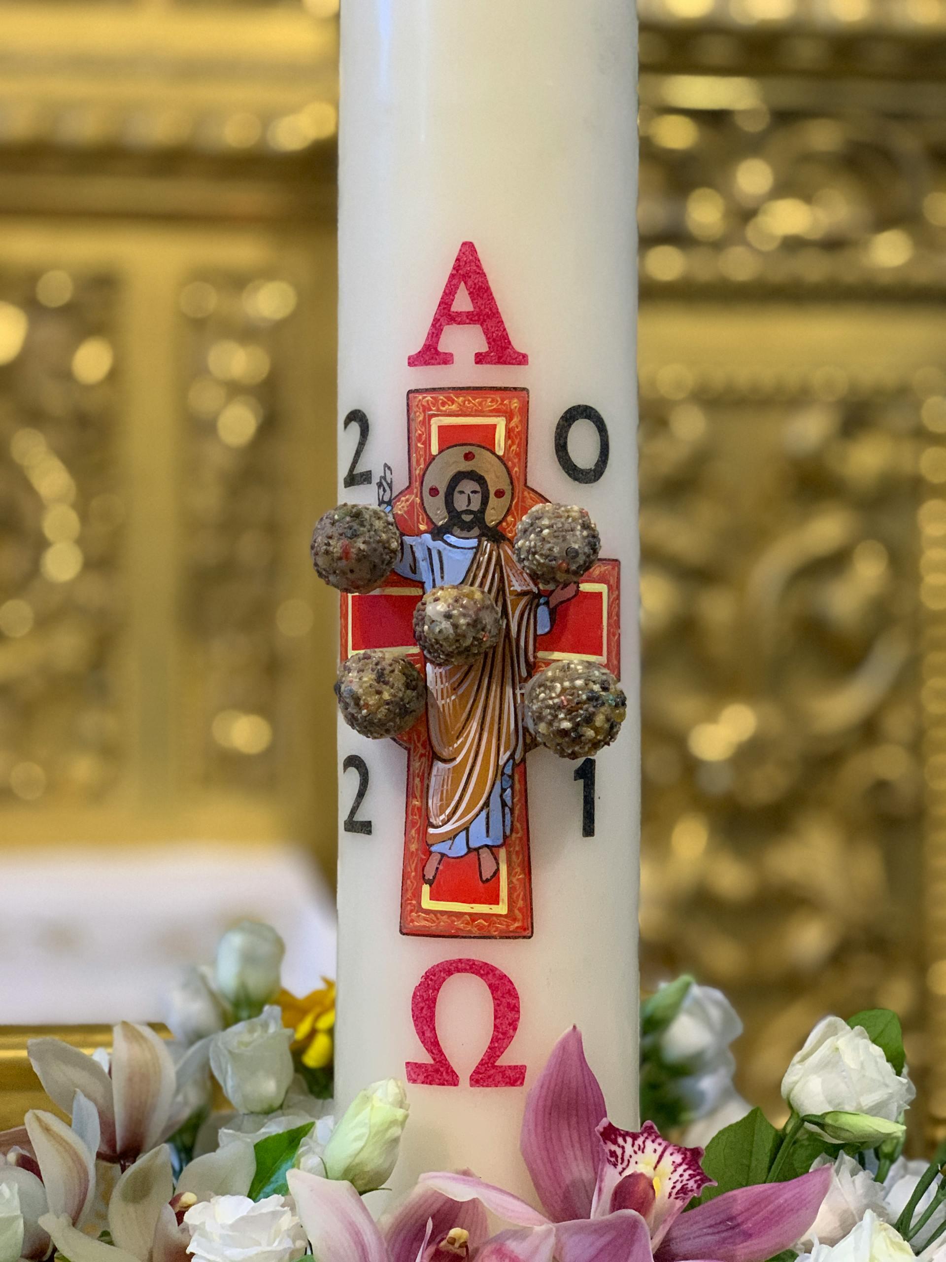 Close-up of a Paschal candle featuring colorful carvings of the required symbols plus a depiction of Jesus on the cross. Five beads of incense are poked into the candle around Jesus.