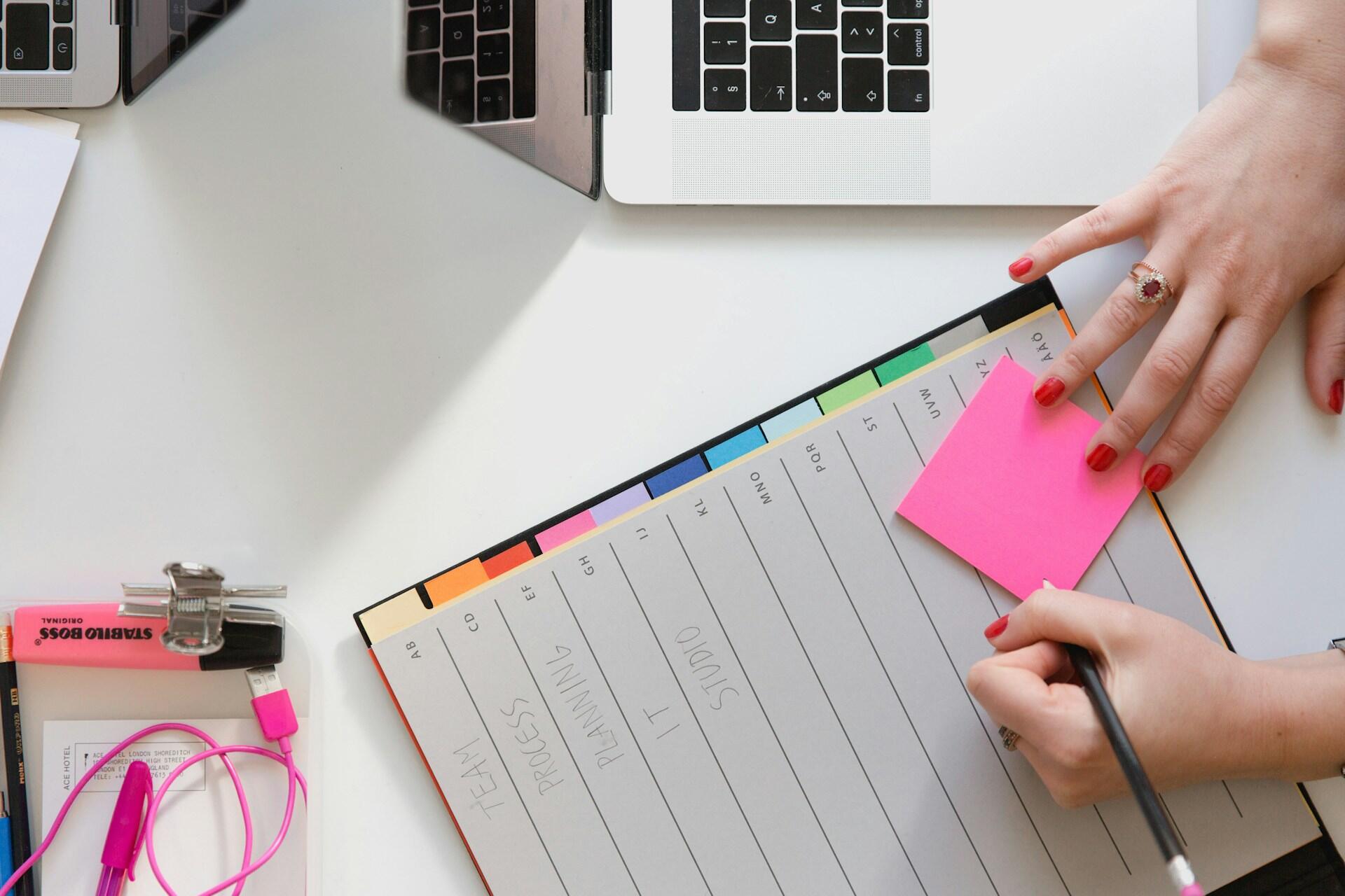 A person writes on a pink sticky note next to a computer.