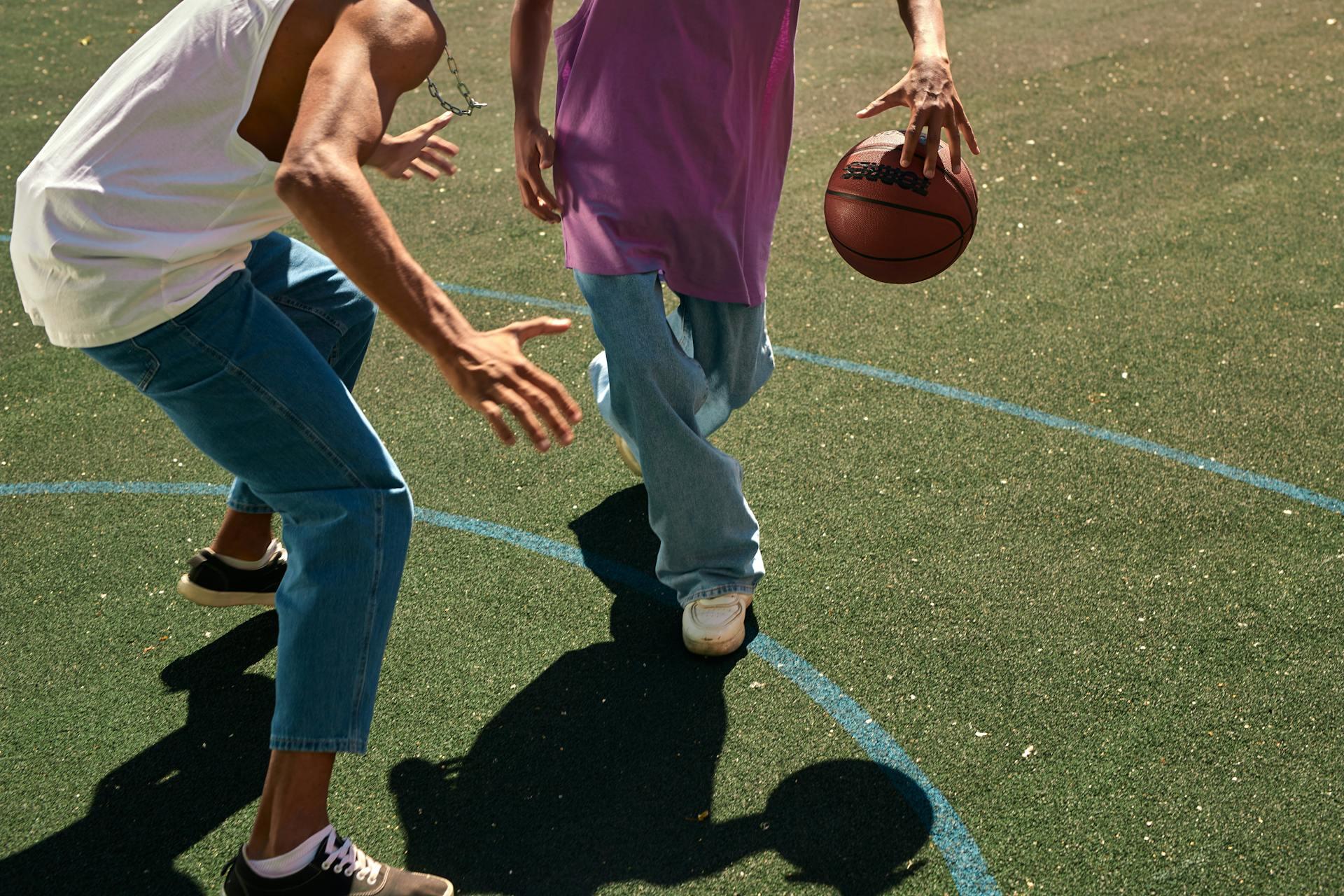 Two individuals on a basketball court, one dribbling a ball, while the other reaches towards it, casting shadows on the ground.