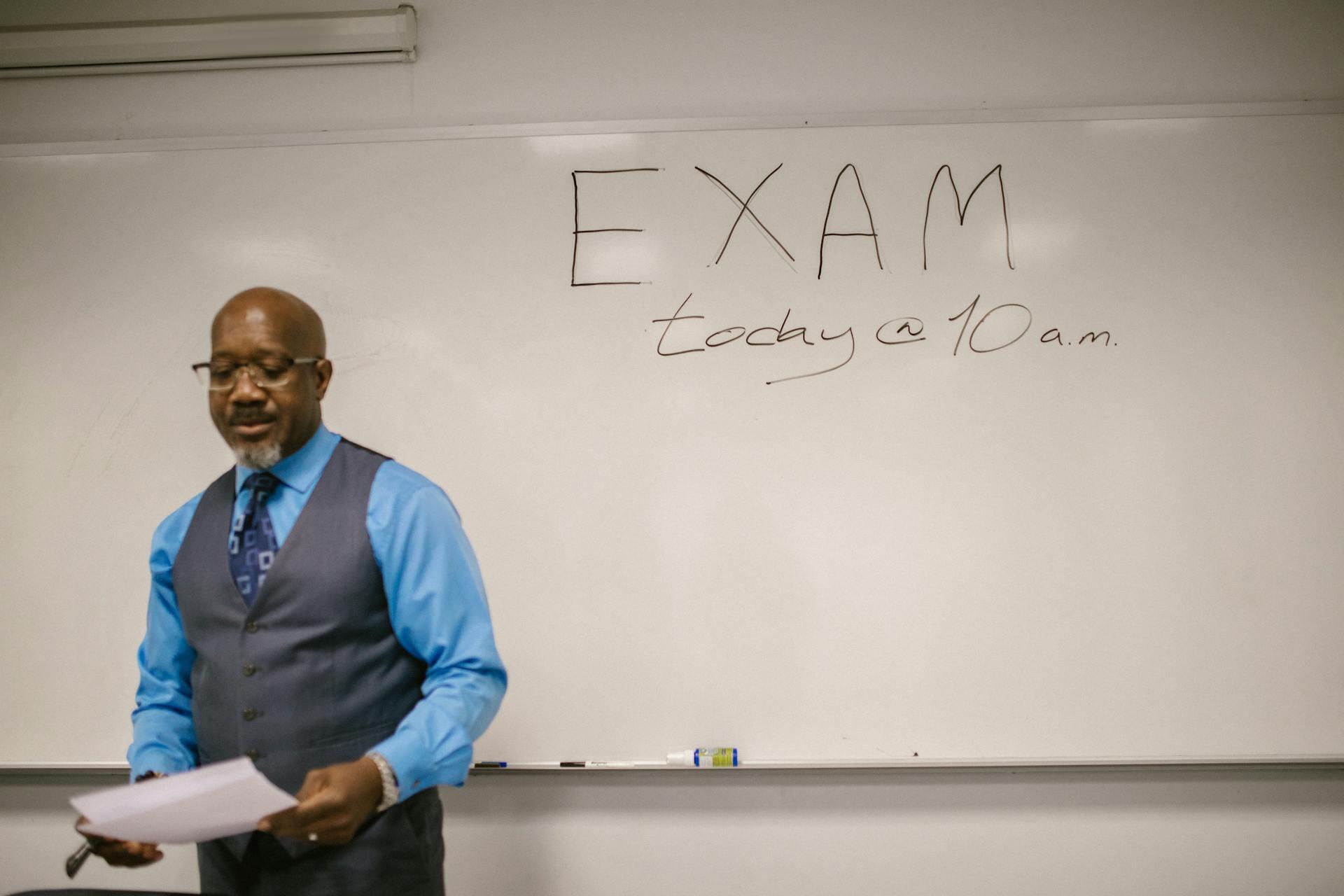 A person in a blue shirt and gray vest stands in front of a whiteboard that reads "EXAM today @ 10 a.m." with papers in hand.