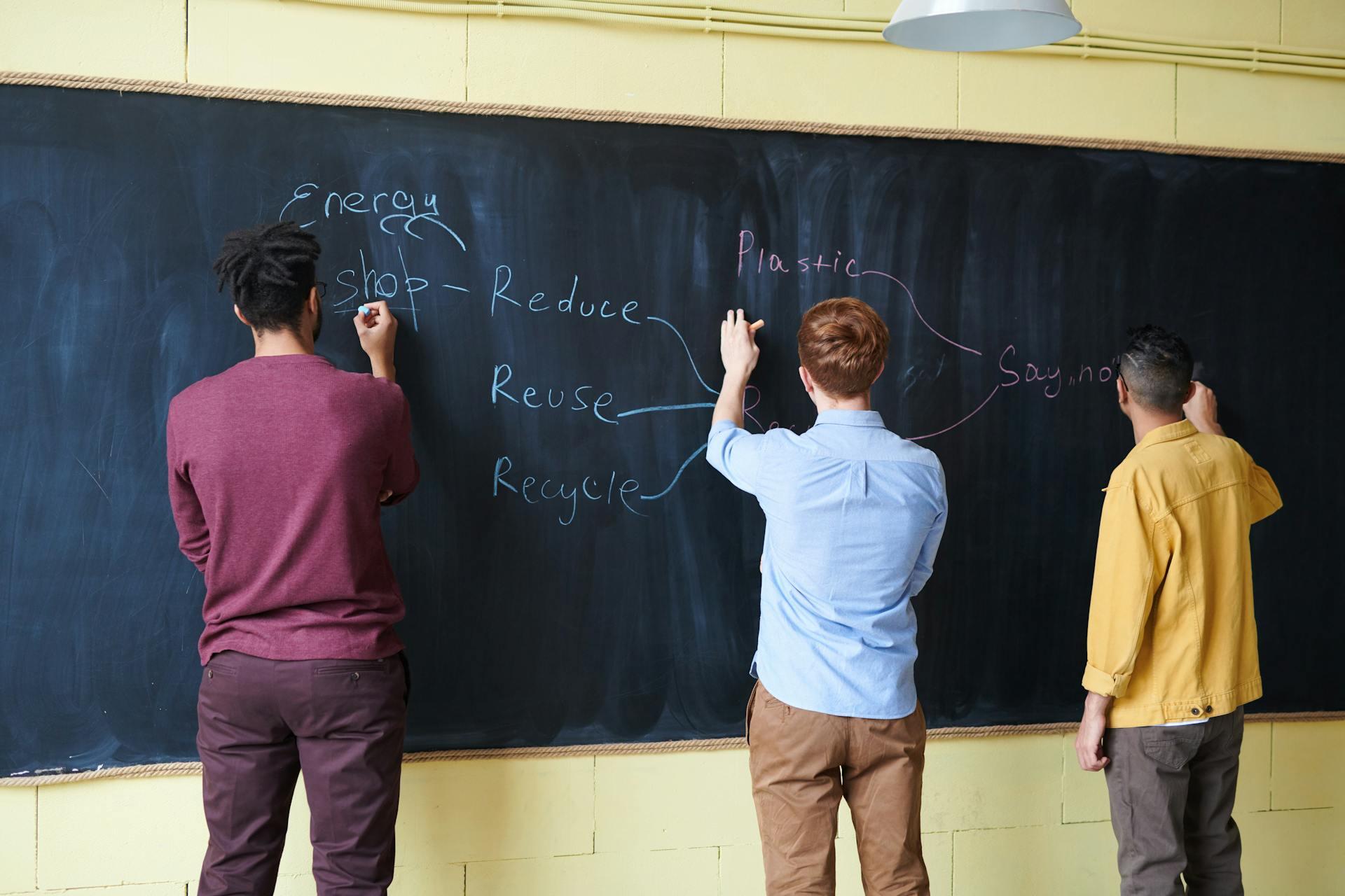 Three people are writing on a chalkboard, discussing topics like "Reduce, Reuse, Recycle" and "Energy" while collaborating in a classroom setting.