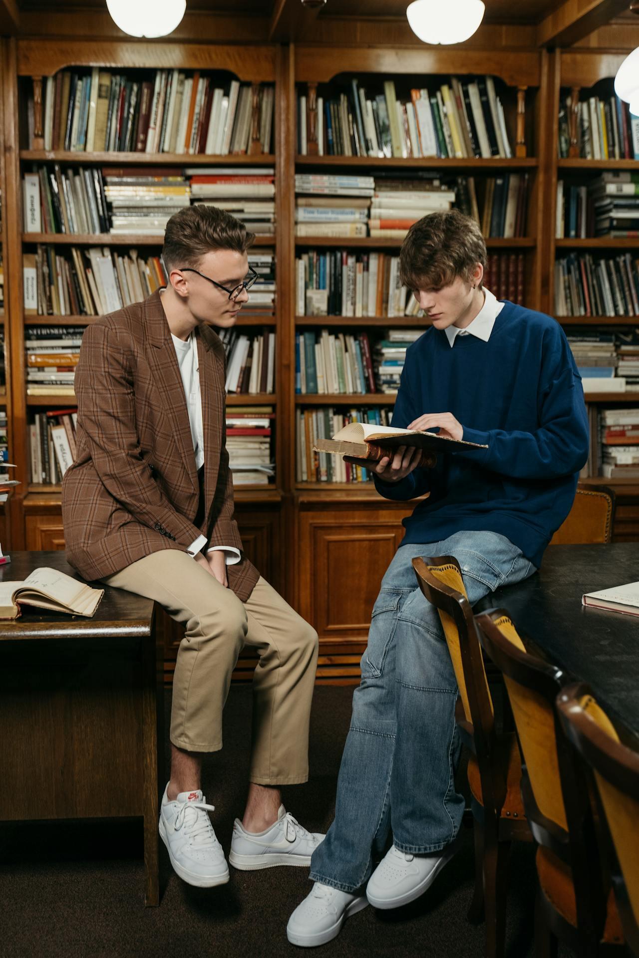 Two young men are seated in a cozy library, discussing a book amid shelves filled with various volumes, creating an inviting atmosphere.