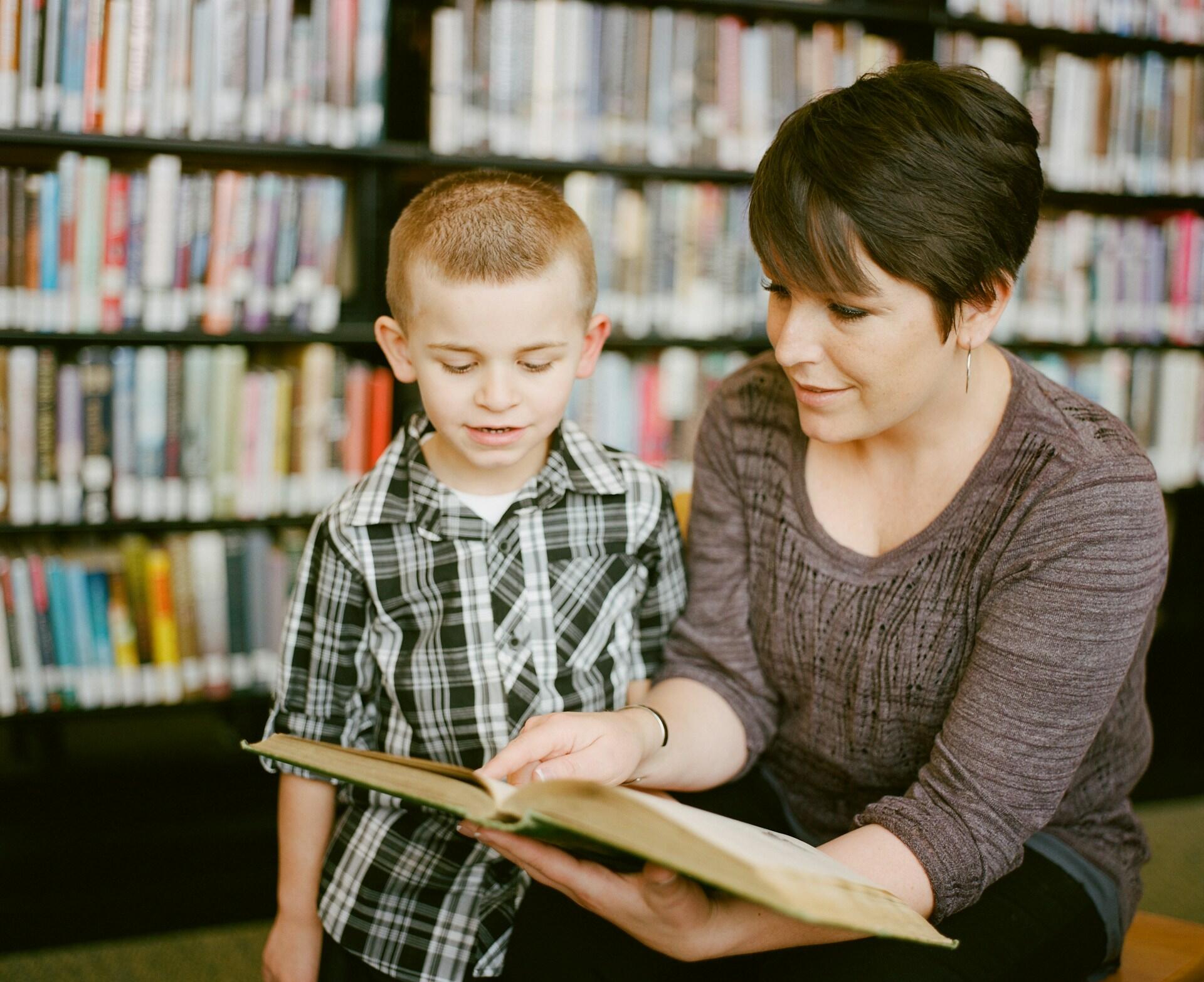 A seated woman with short hair holds a book so the child next to her can see its pages.