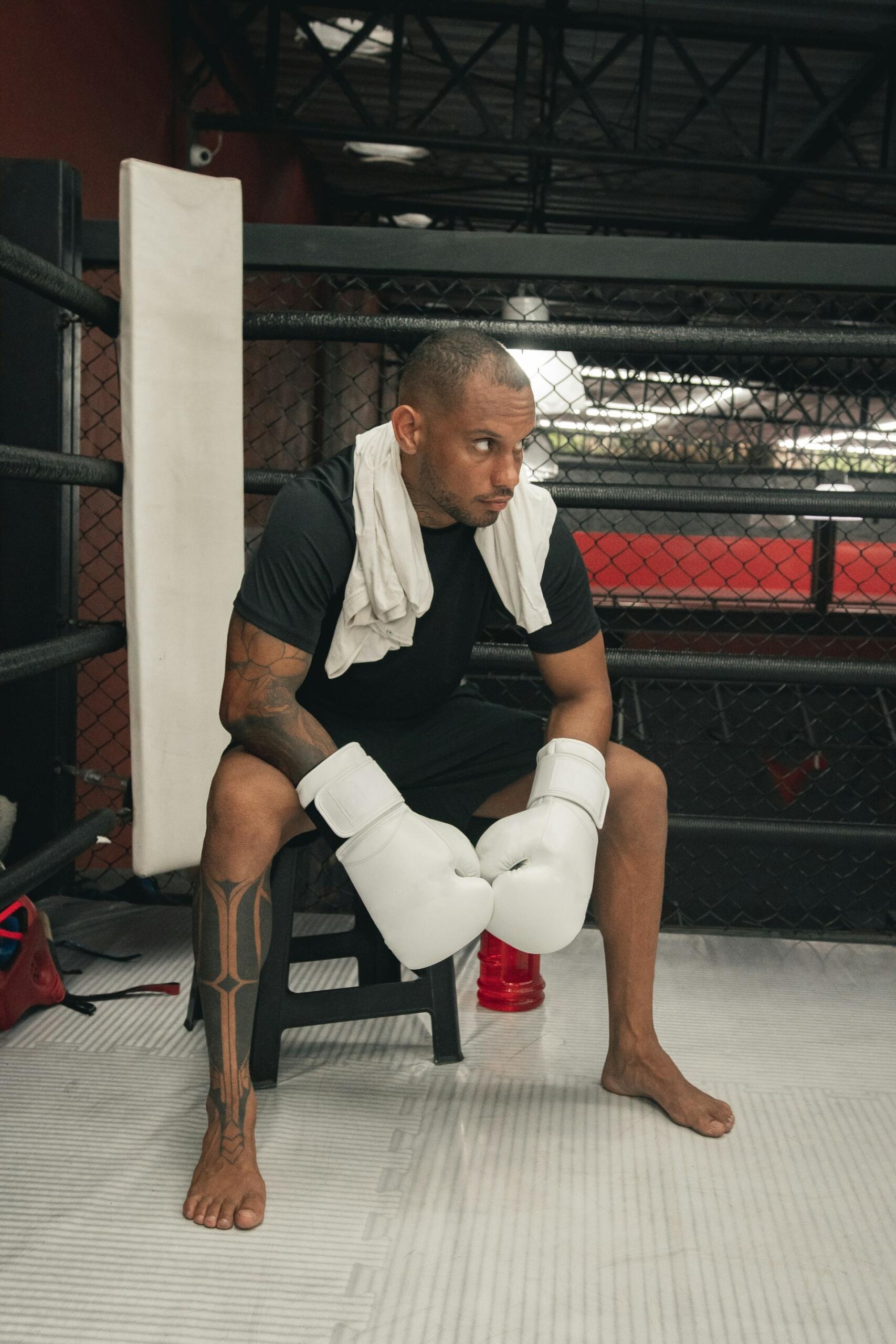 A person wearing boxing gloves sits on a stool outside a boxing ring. 